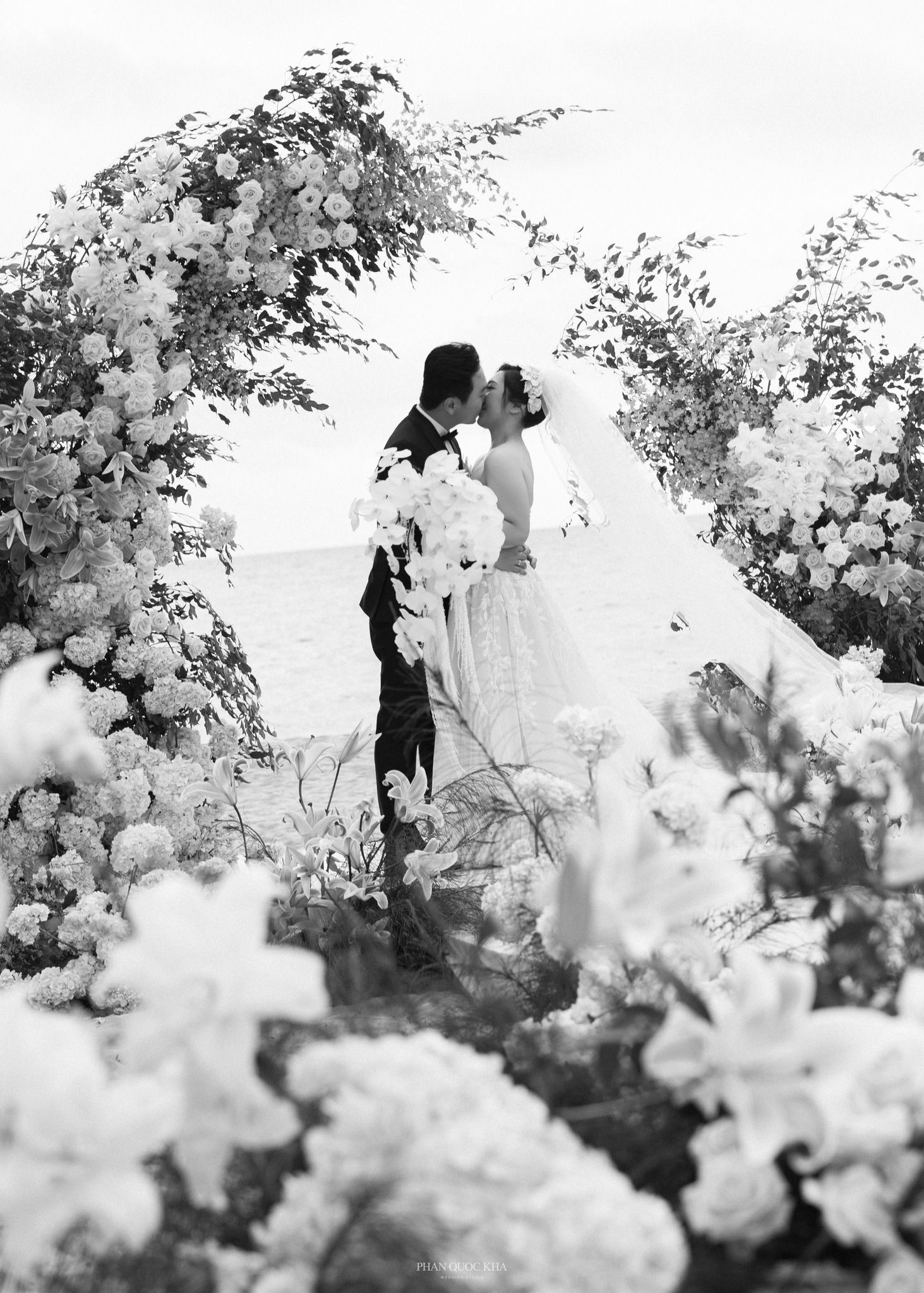 Bride and groom share a kiss under a floral arch surrounded by white blooms at outdoor ceremony.