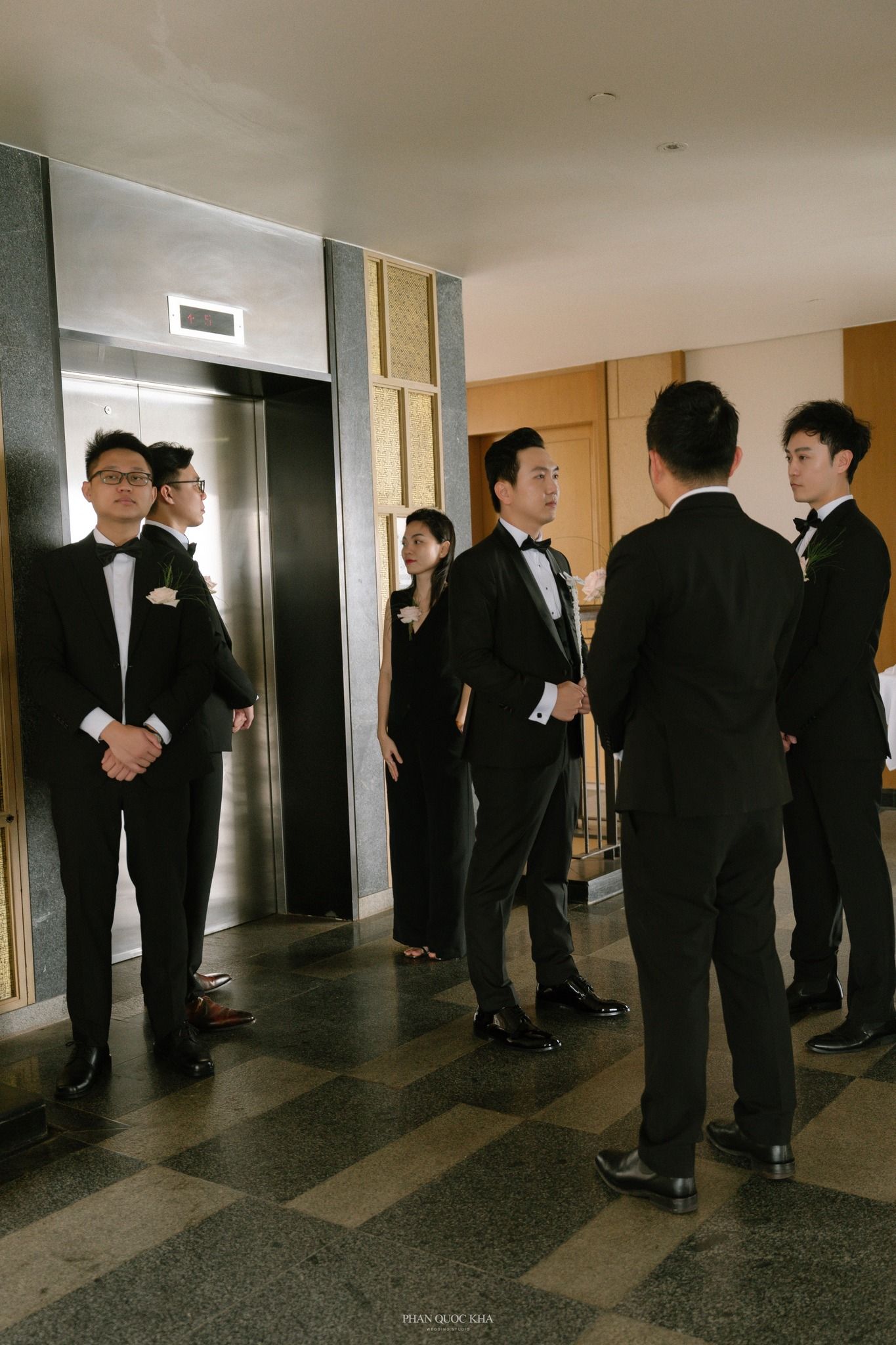 Groomsmen in black tuxedos gather near elevator in hotel lobby before wedding ceremony.