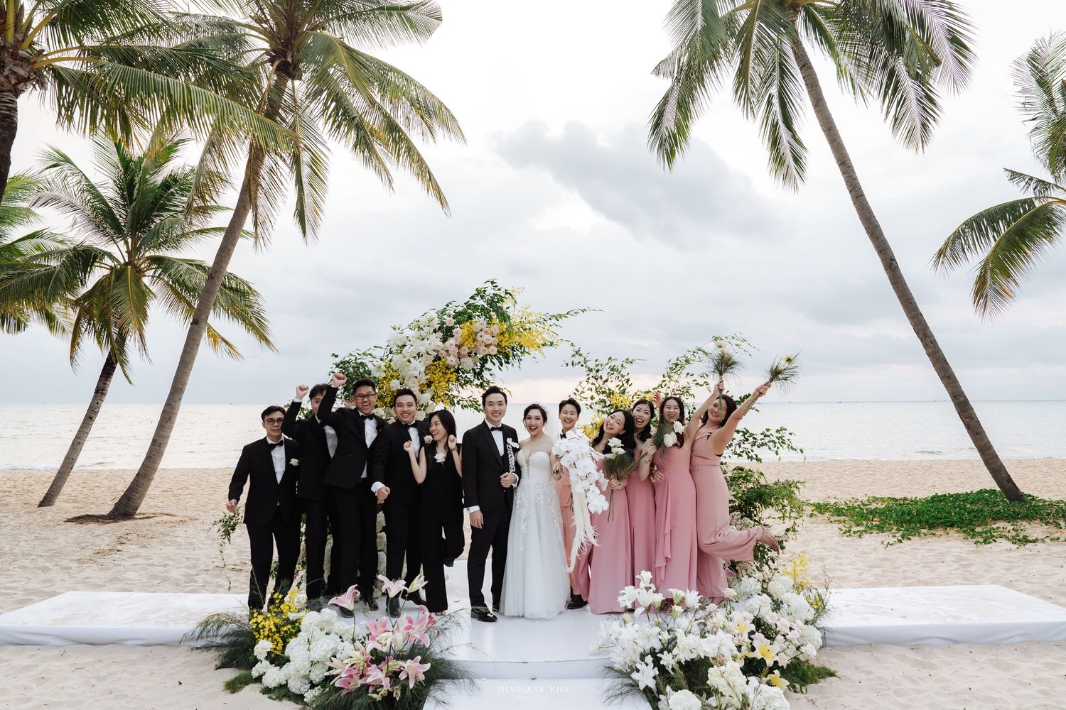 Wedding party poses at beach ceremony with palm trees, floral arch, and bridesmaids in pink dresses.