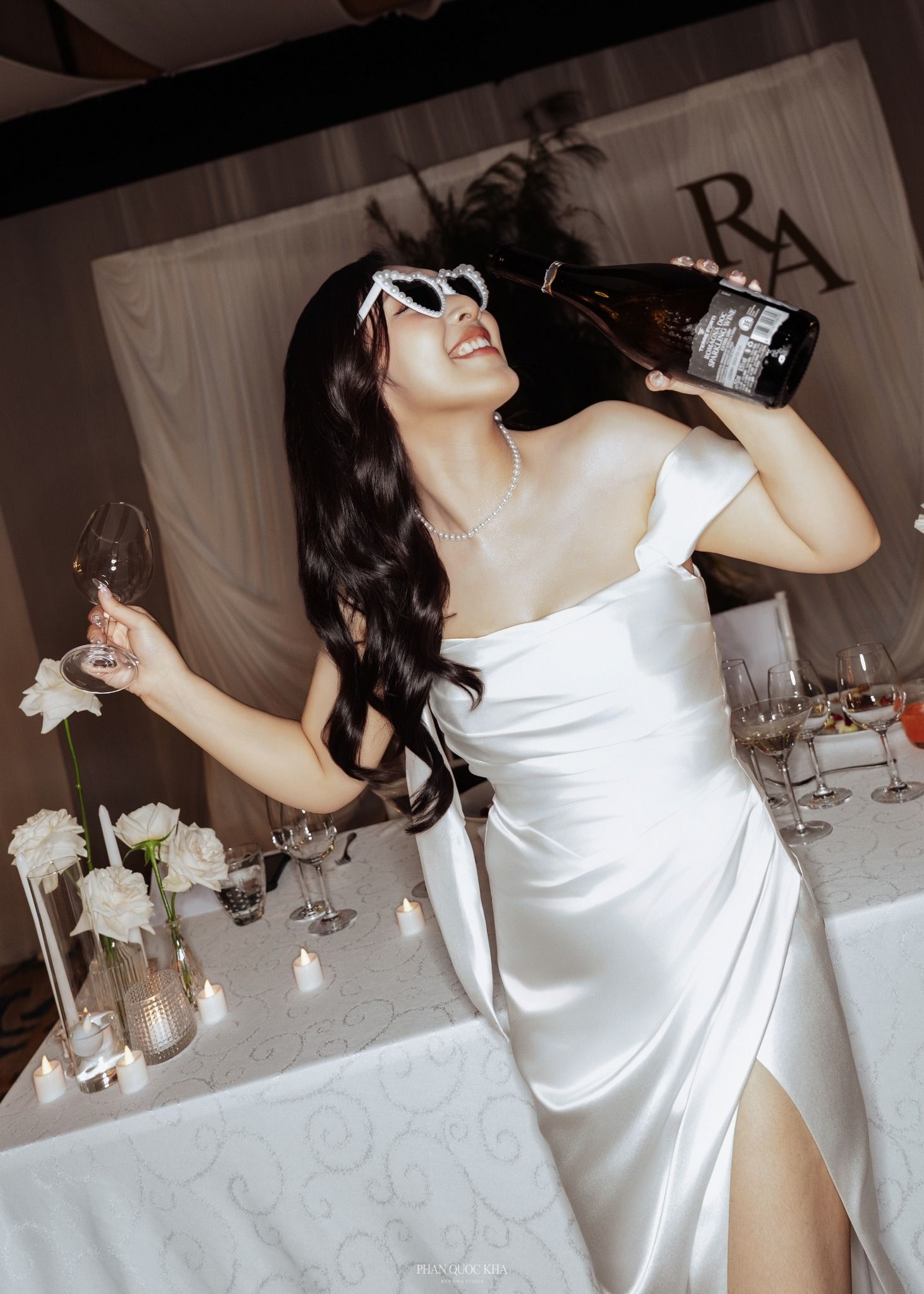 Joyful bride in white satin dress playfully drinks from a champagne bottle at wedding reception.