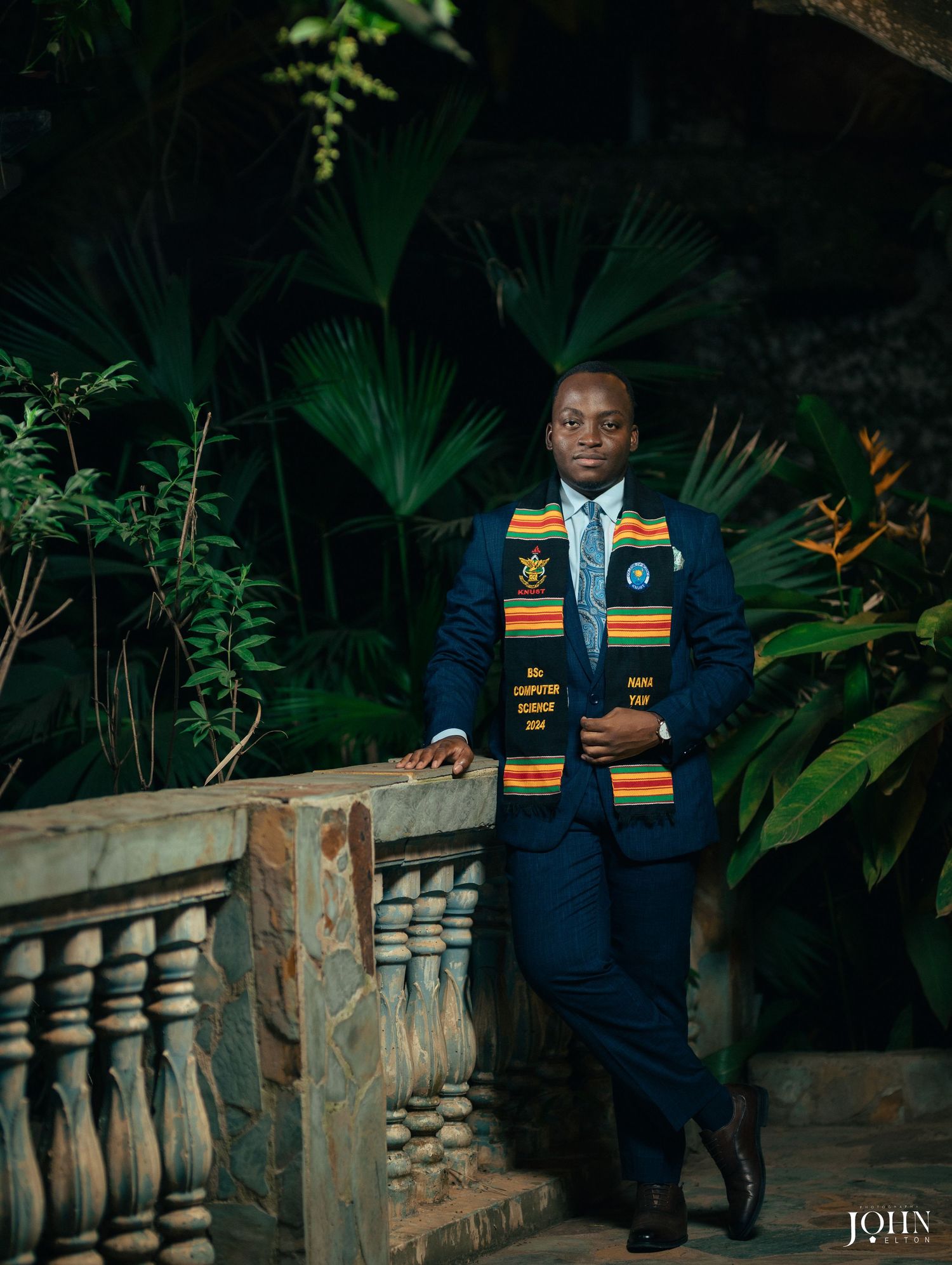 Graduate student in cap and gown poses confidently against modern interior railing with ambient lighting.