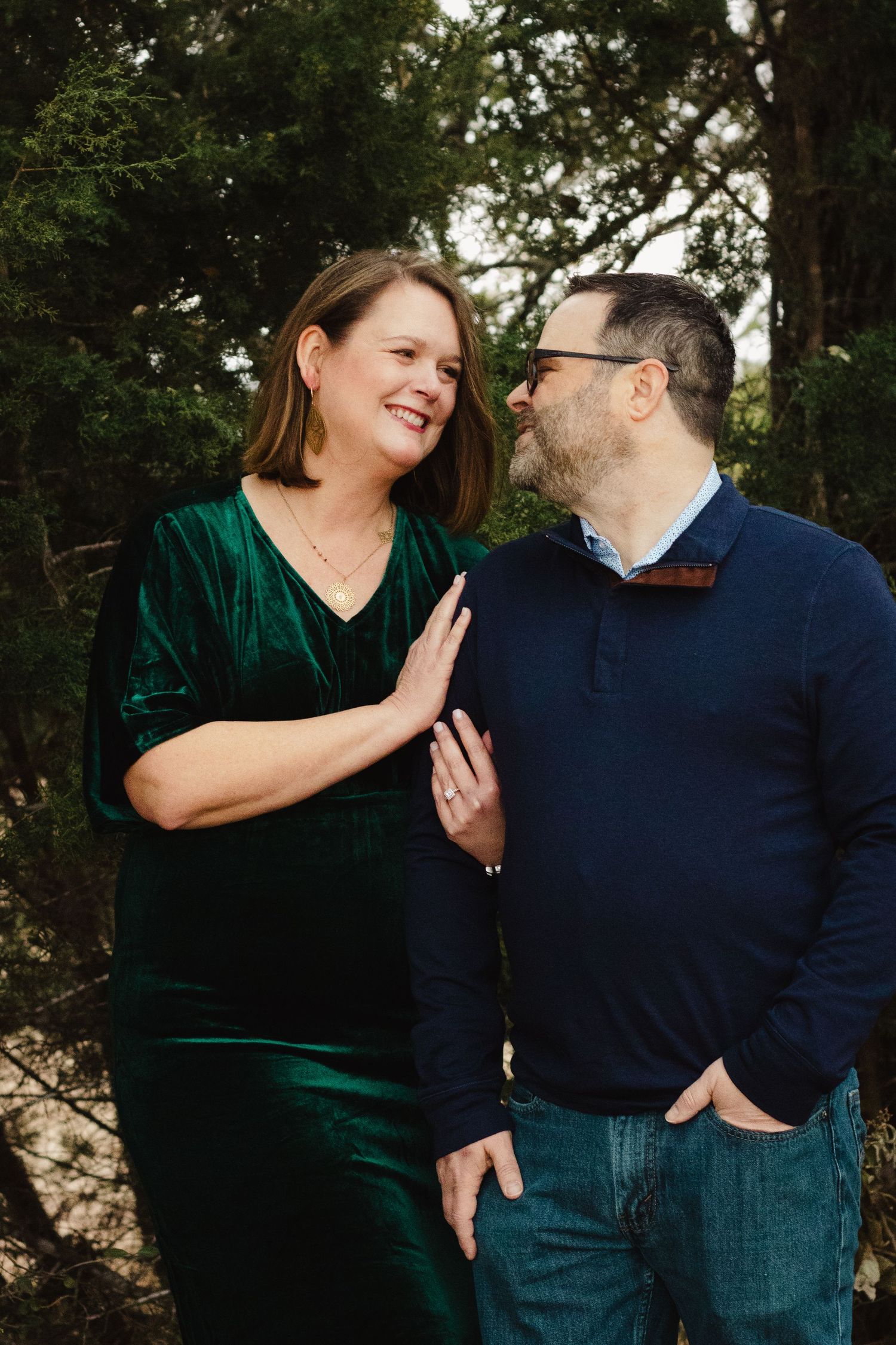 A couple shares a playful moment outdoors with one wearing a green velvet top and the other in a navy sweater.