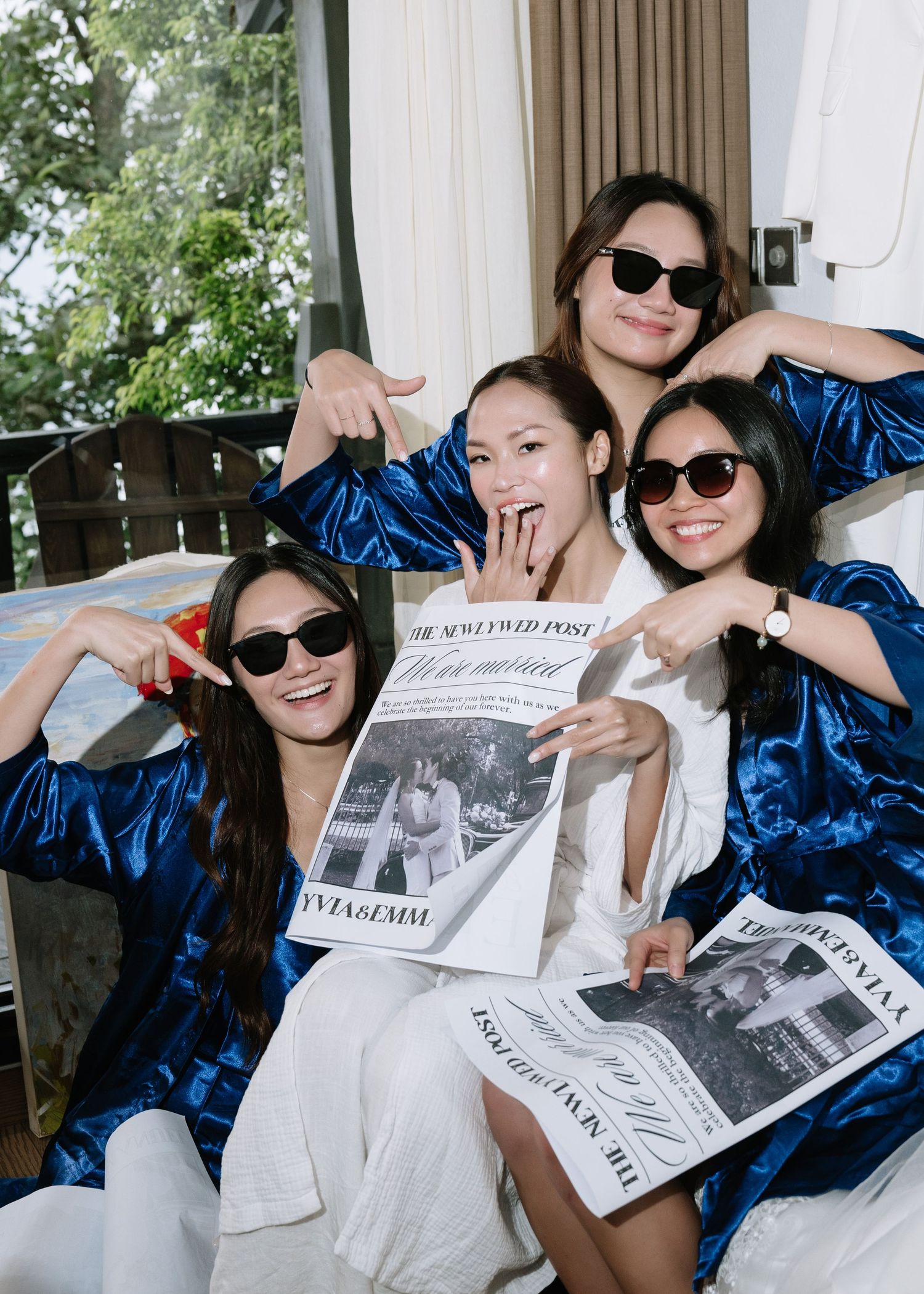 Four women in blue robes and sunglasses pose with a wedding newspaper at a bridal party celebration.