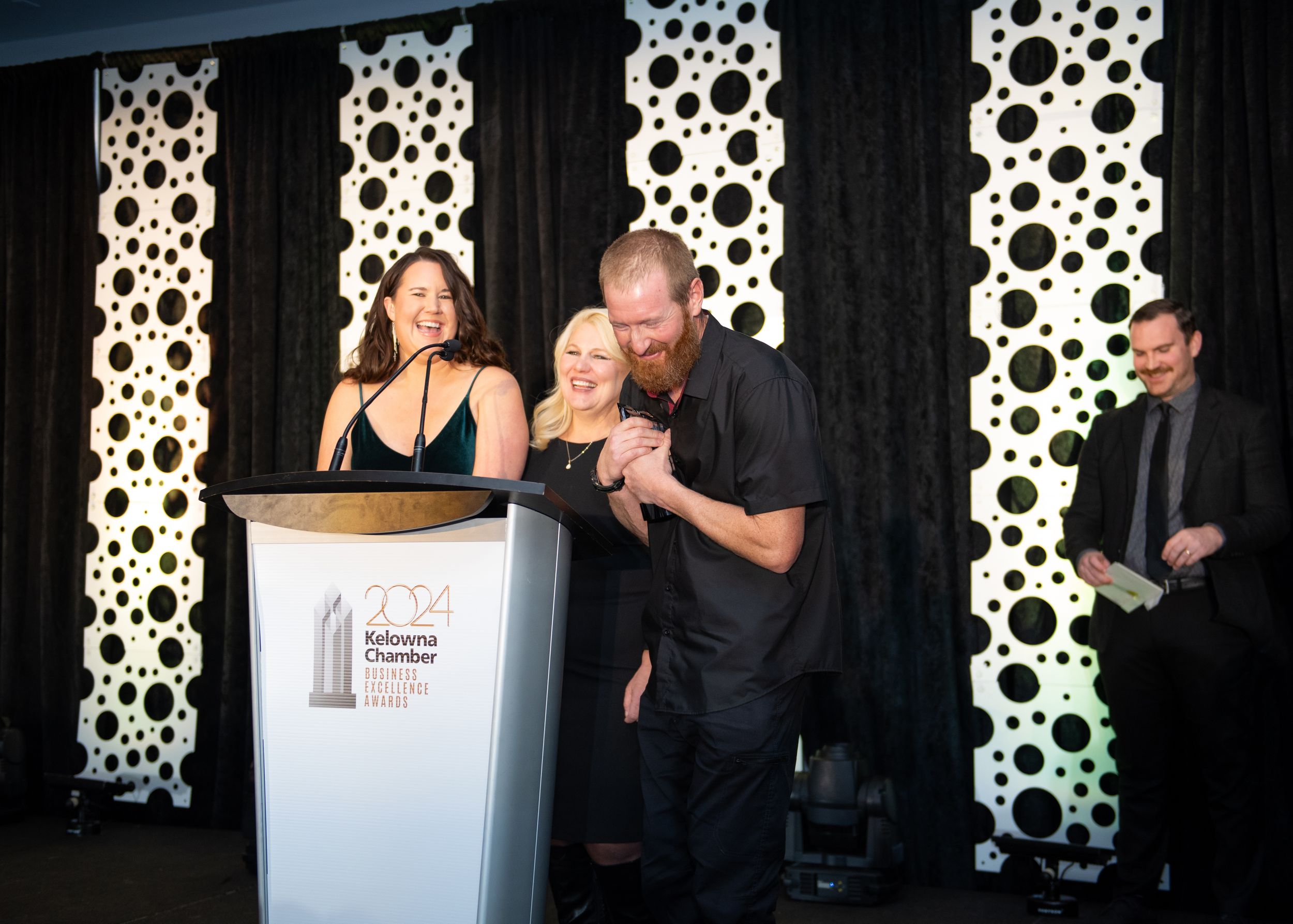 Man with red beard in black outfit celebrating on stage after receiving award at Kelowna Chamber Business Excellence Awards.