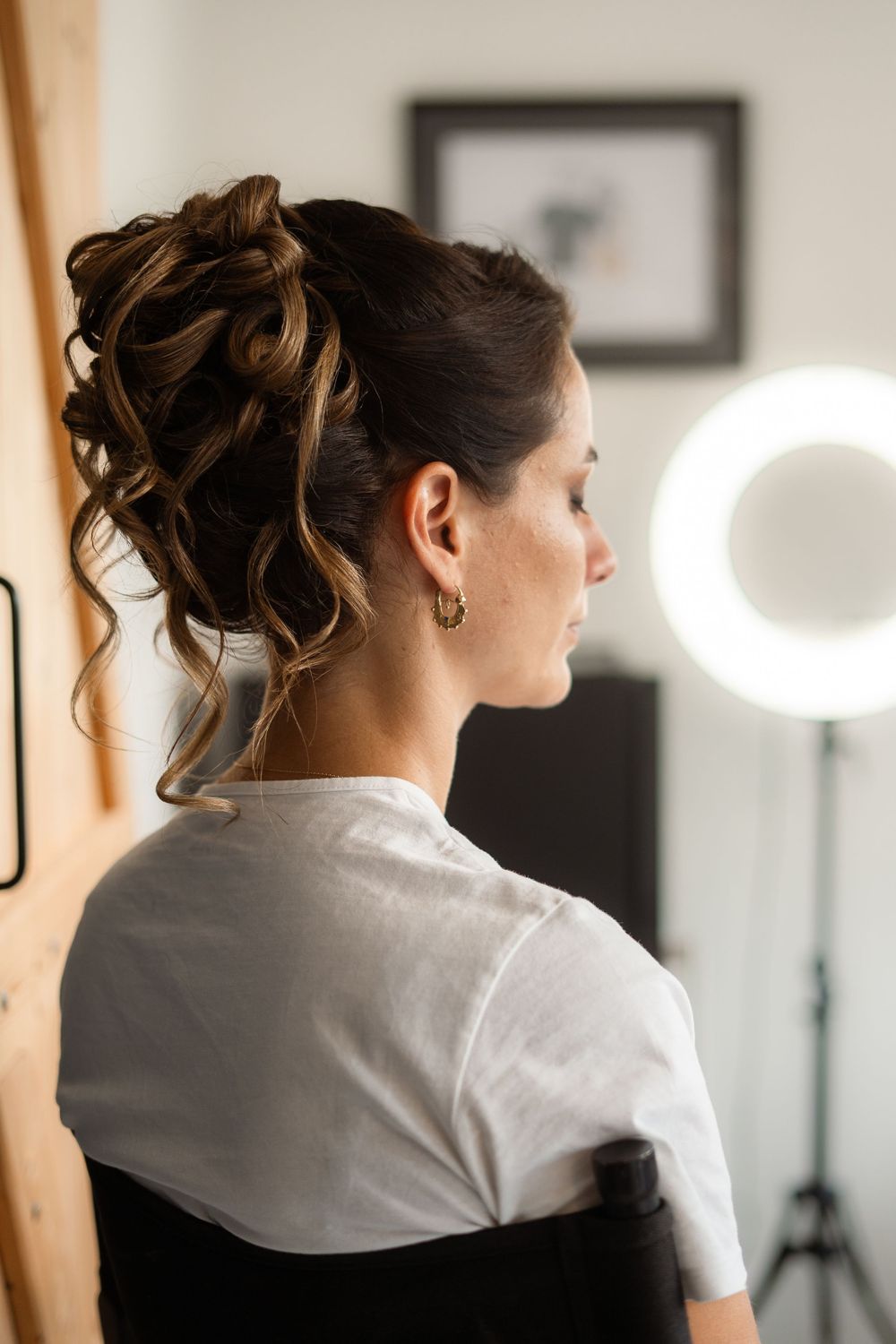 A woman in a white dress getting ready with elegant hair and makeup in a natural lit room.