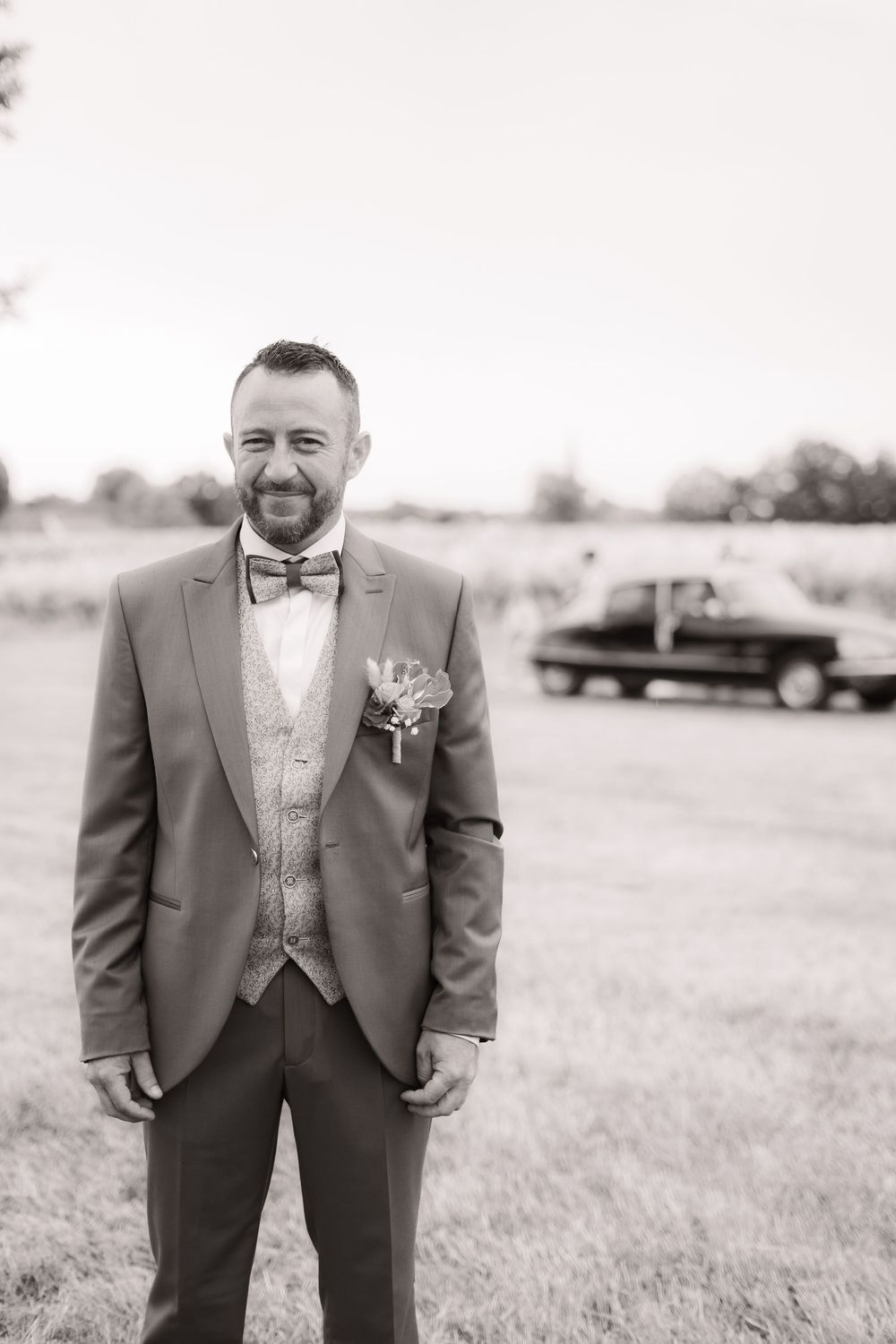 Groom in a classic charcoal suit with bow tie standing in an outdoor field setting.