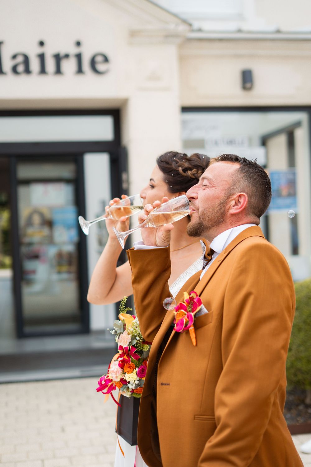 Wedding party in brown suits and white dress drinking champagne outside a town hall building.