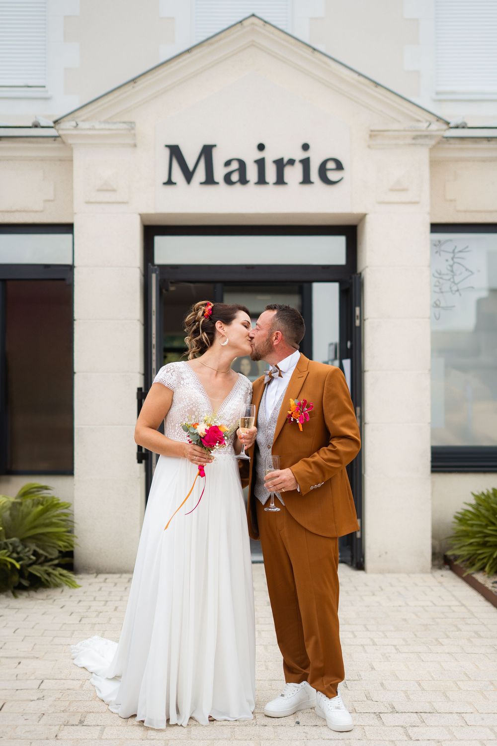 Newlyweds in white dress and brown suit sharing a celebratory moment outside a Mairie building.