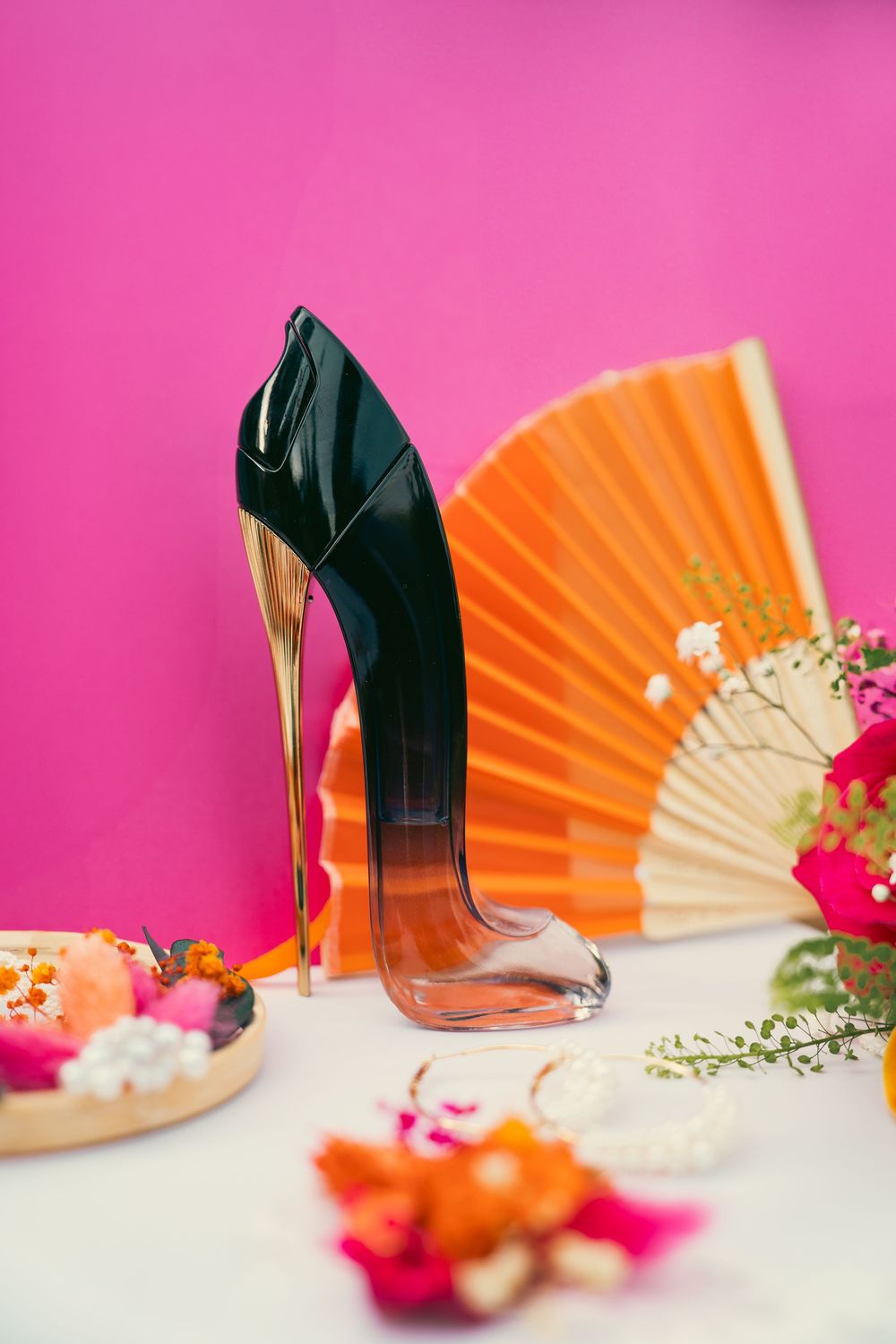Elegant black high heels displayed with orange fans and pink flowers against a vibrant pink background.