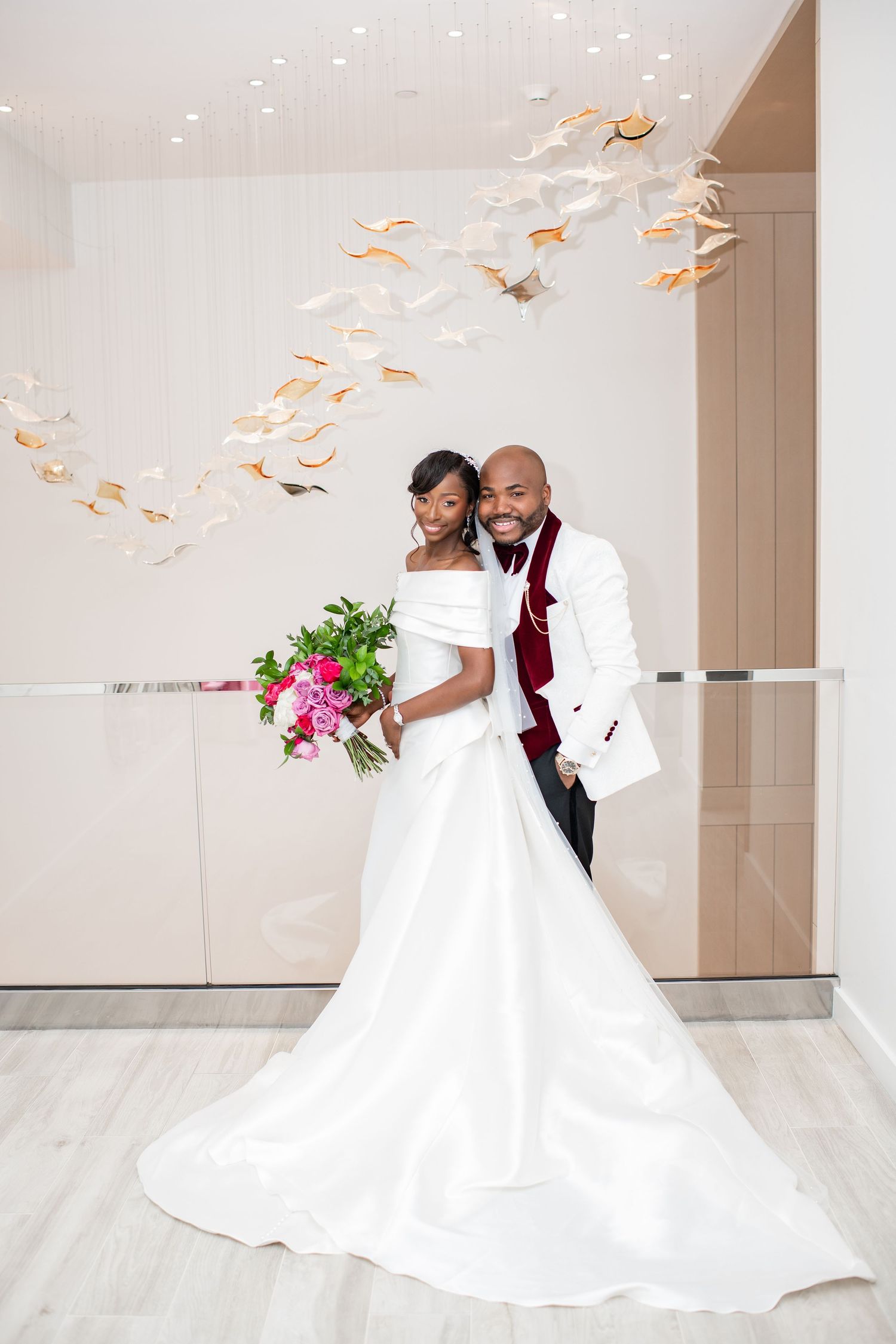 Bride in white off-shoulder gown and groom in burgundy vest pose in a modern wedding venue.