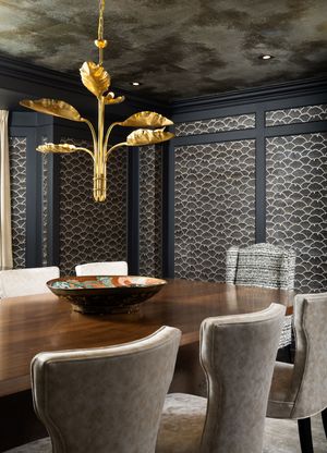 Modern dining room with gold ginkgo leaf chandelier, dark walls, and velvet upholstered chairs around wooden table.