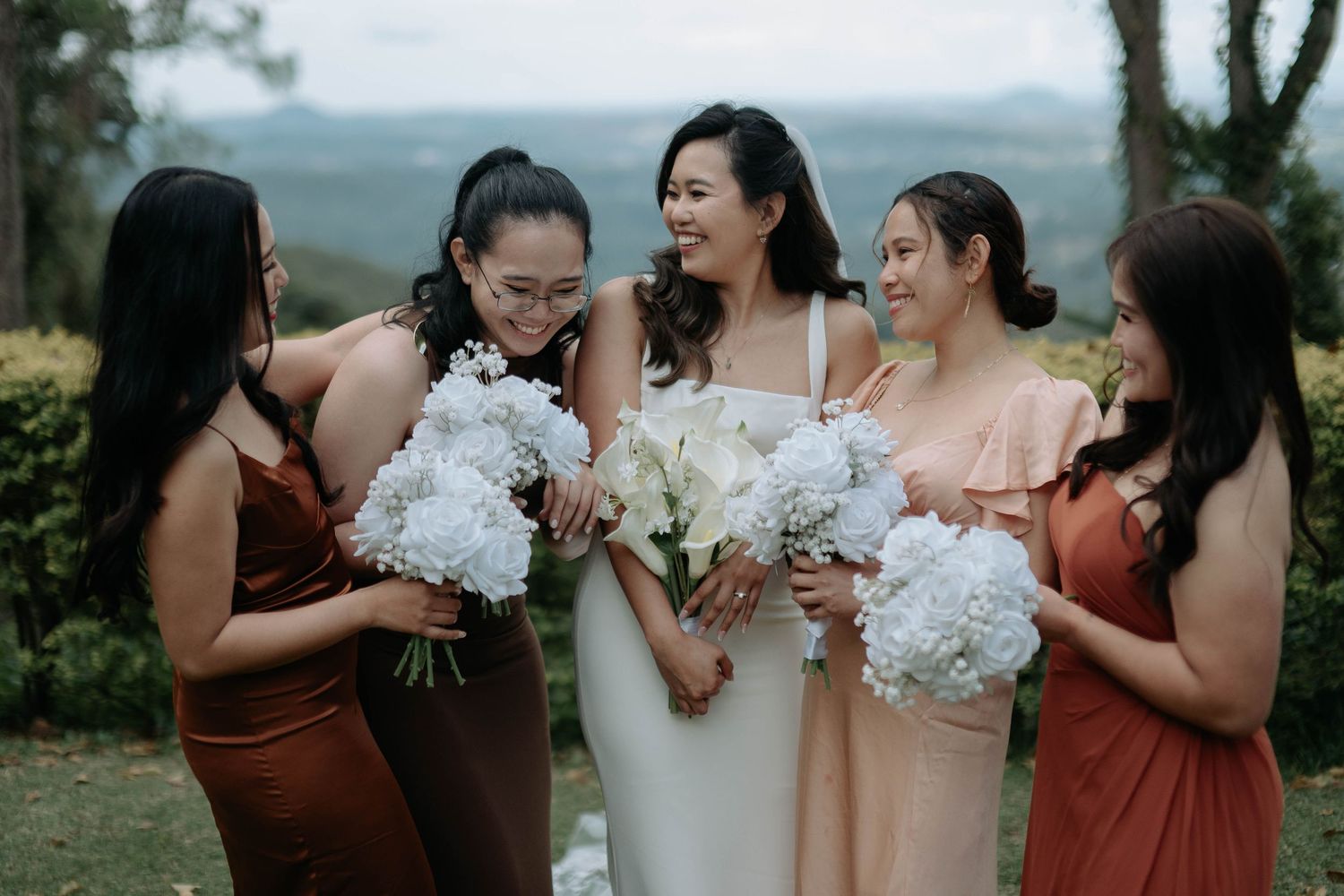 A wedding party in rust-colored and blush dresses stands together holding white floral bouquets in an outdoor setting.
