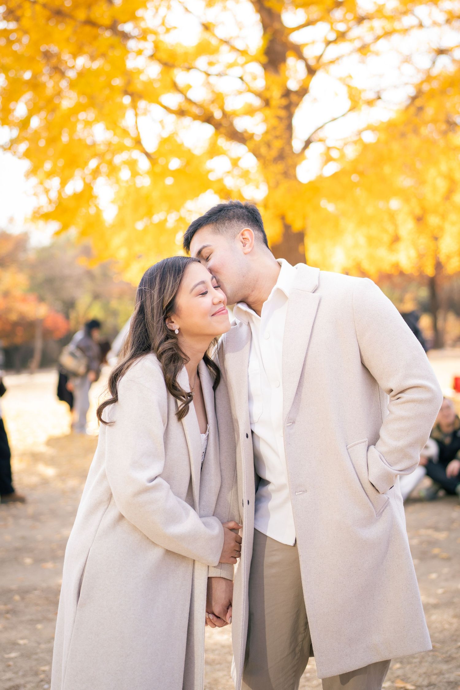 A romantic couple in matching beige coats share an intimate moment under golden autumn foliage in a park.