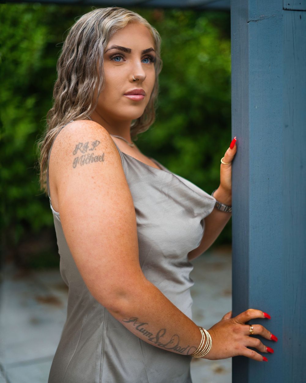 Woman in gray dress poses against a blue wall in an outdoor setting with lush greenery in background.