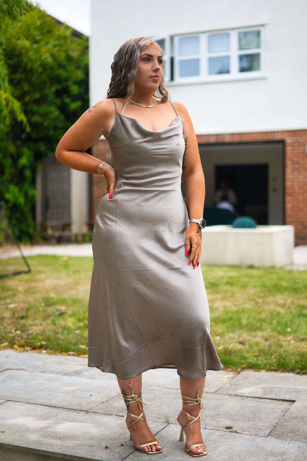 Someone poses in a silky gray slip dress and strappy heels on a paved walkway outside a house.