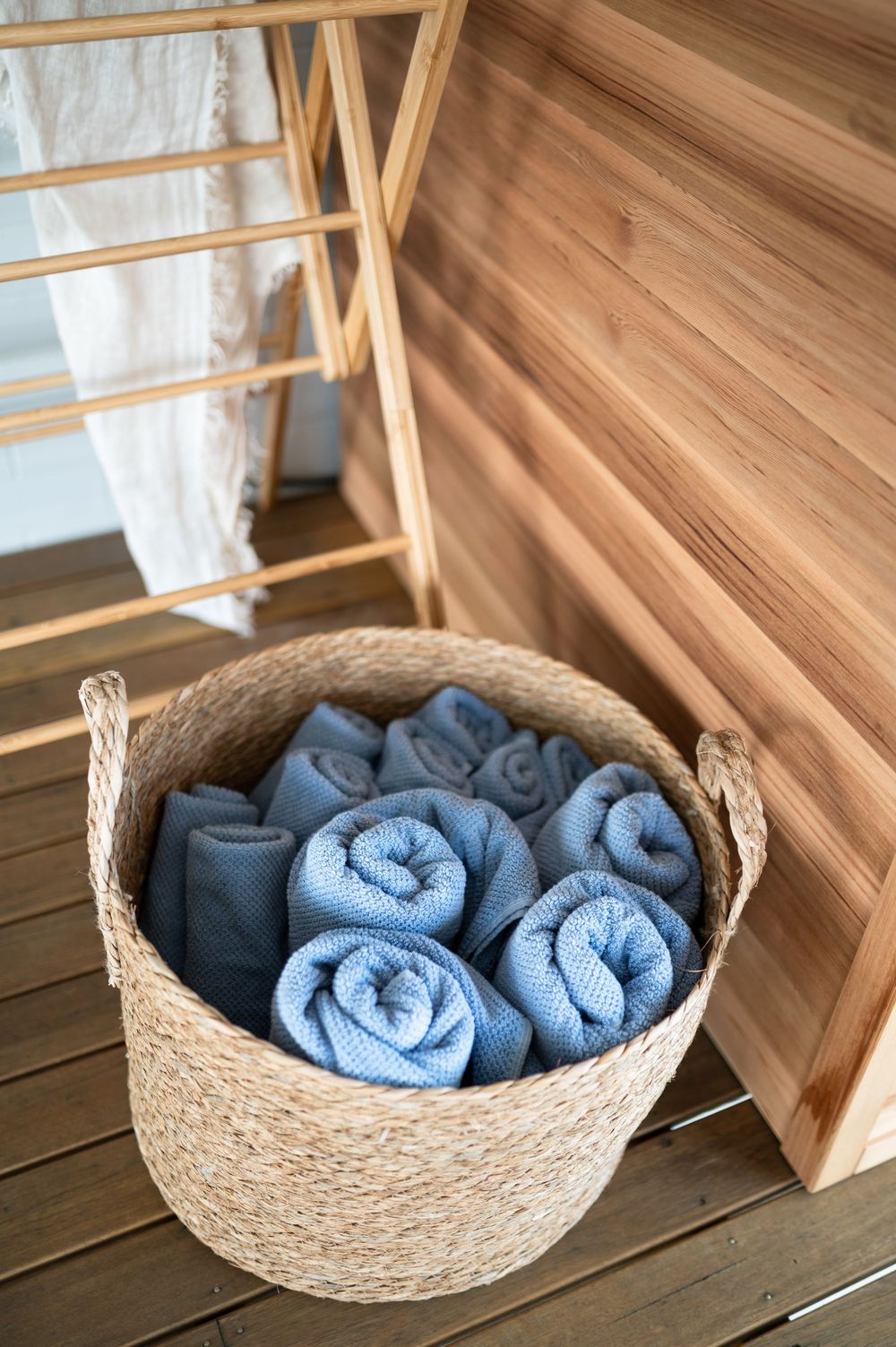 A woven natural seagrass basket filled with rolled blue towels sits on a wooden floor beneath storage shelves.