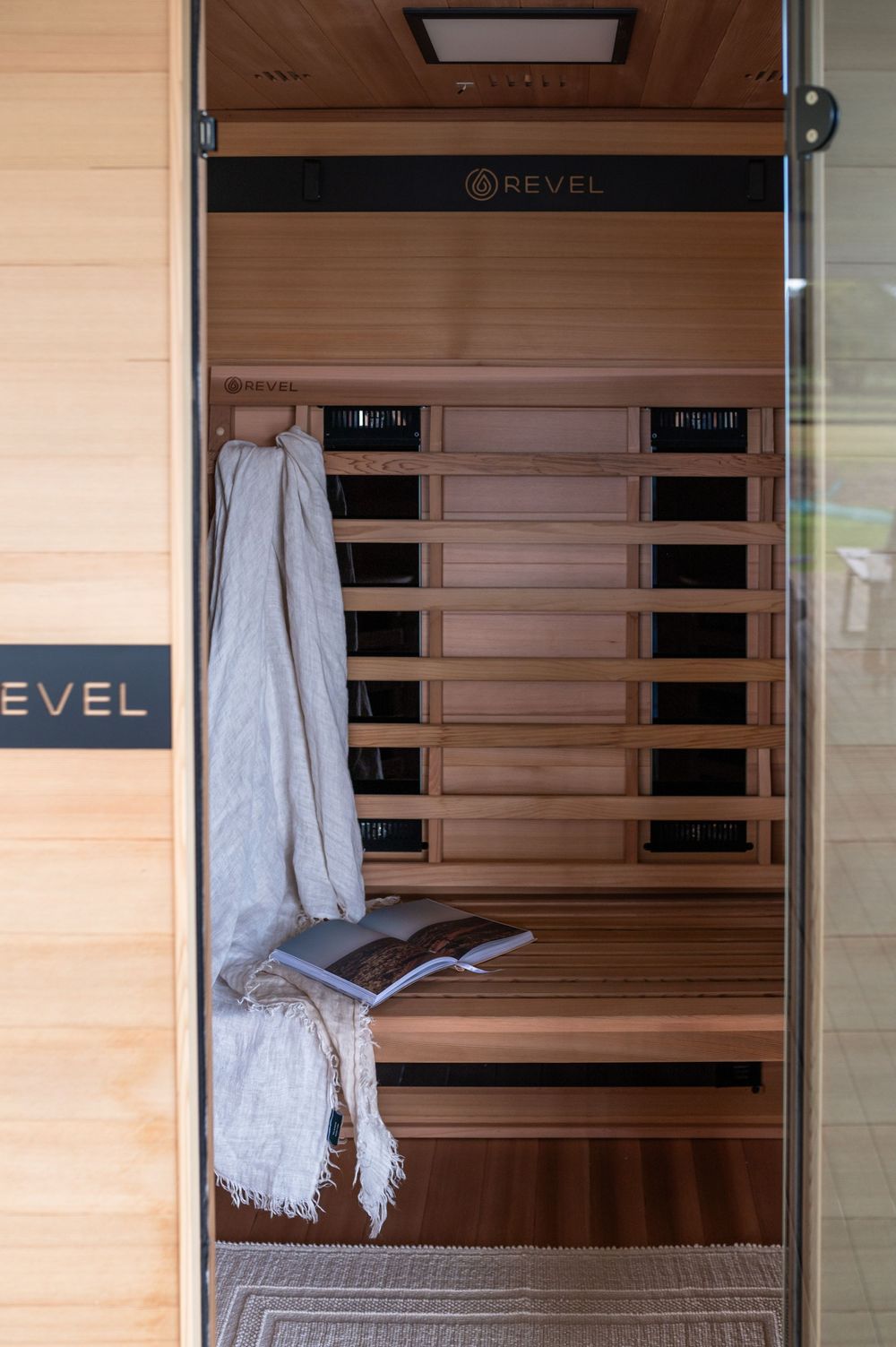 Modern wooden infrared sauna interior with white towel and black digital control panel visible through glass door.