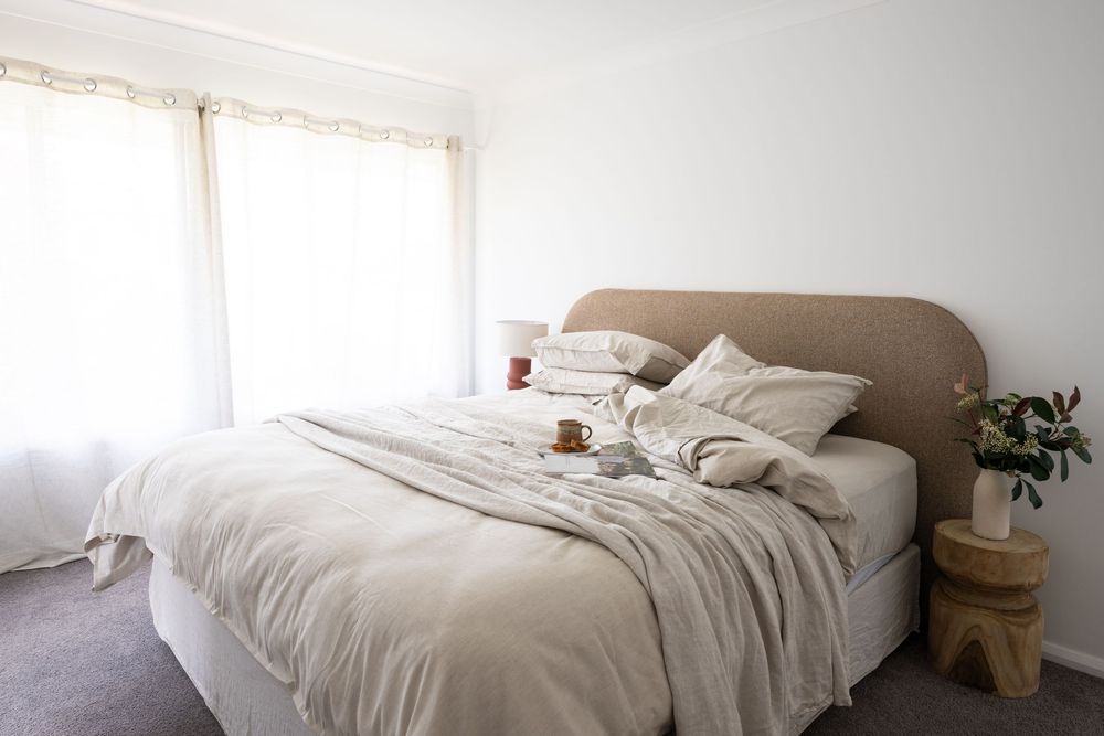 A bright minimalist bedroom with white bedding, natural wood accents, and sunlight streaming through sheer curtains.