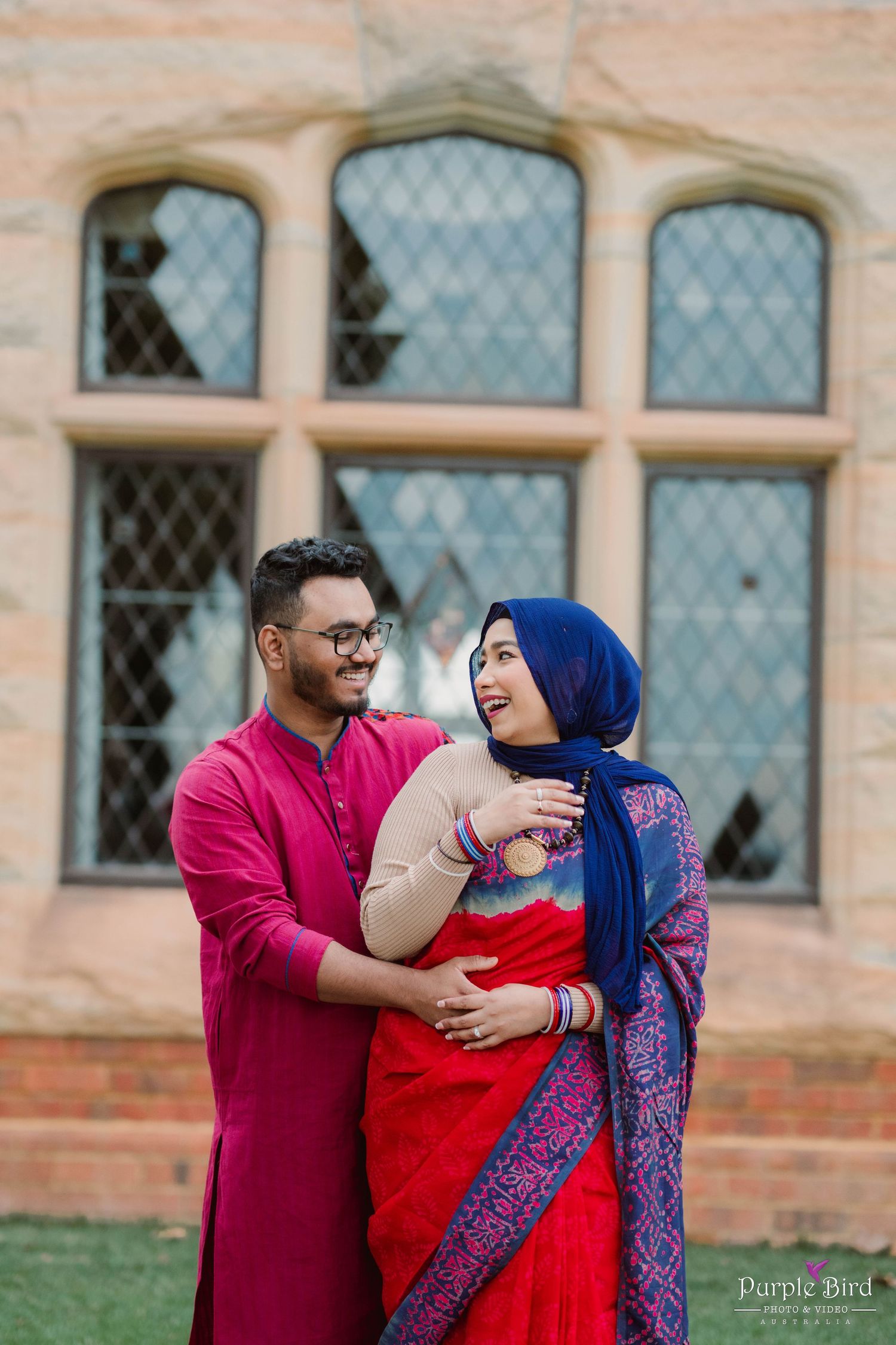 A couple in traditional attire sharing a joyful moment in front of a historic building with diamond-paned windows.
