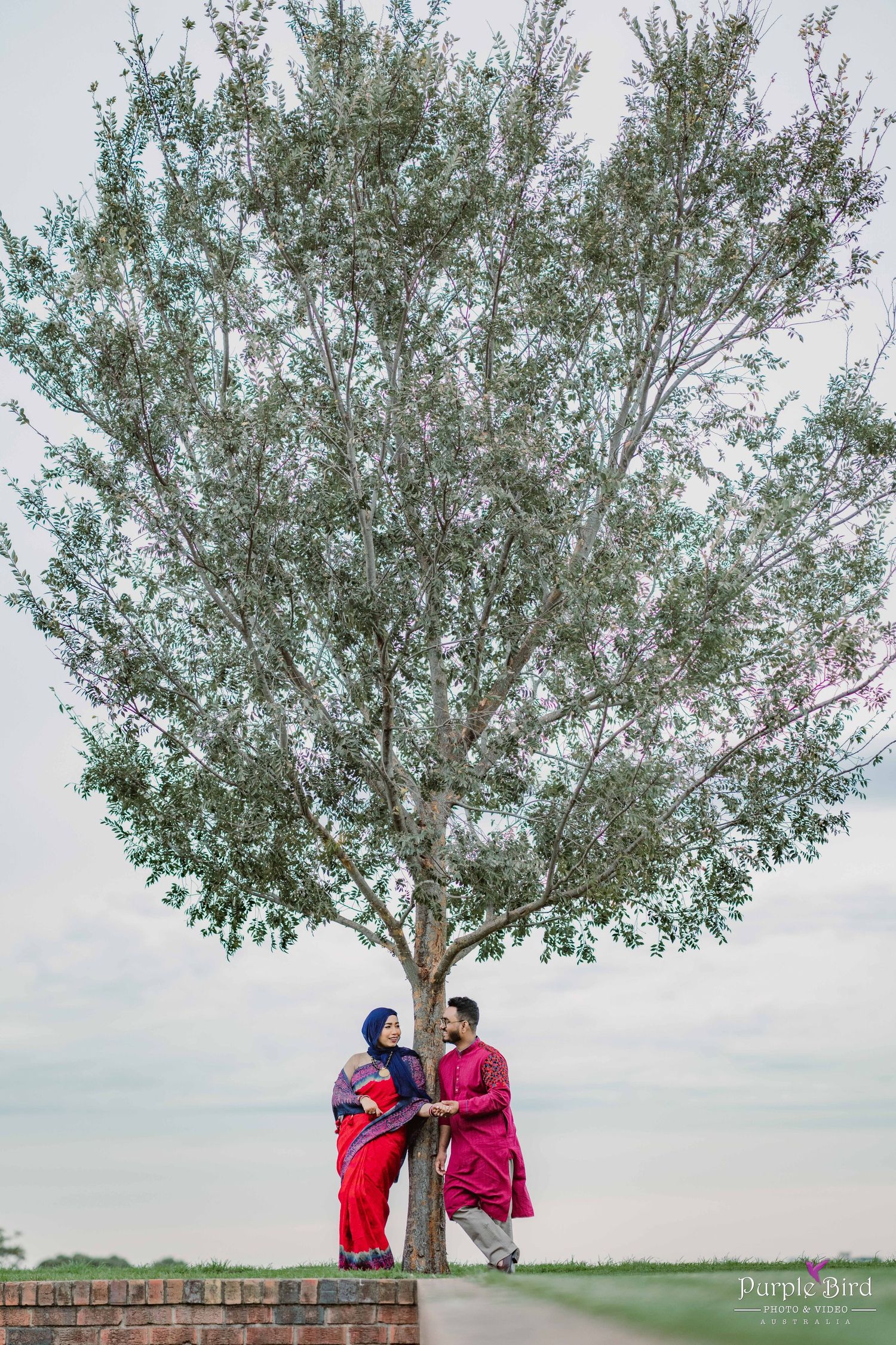 A couple poses beneath a large green tree in an outdoor setting with cloudy skies as backdrop.