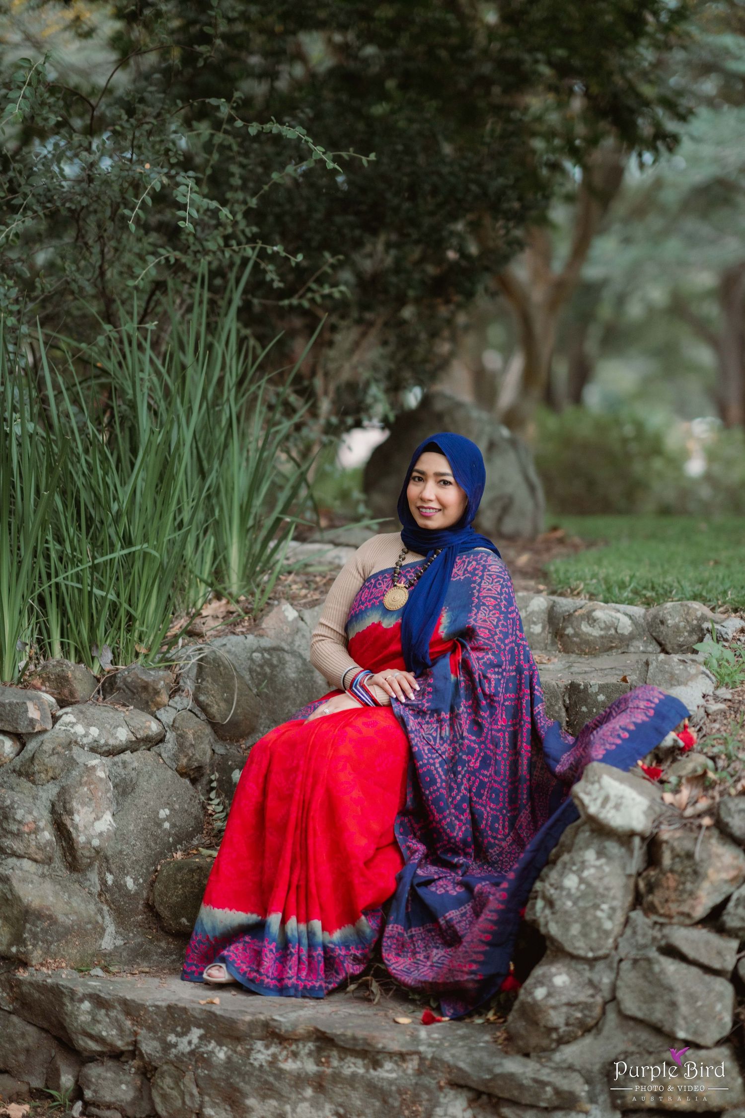 A person in a red and blue sari sits on stone wall surrounded by lush greenery.