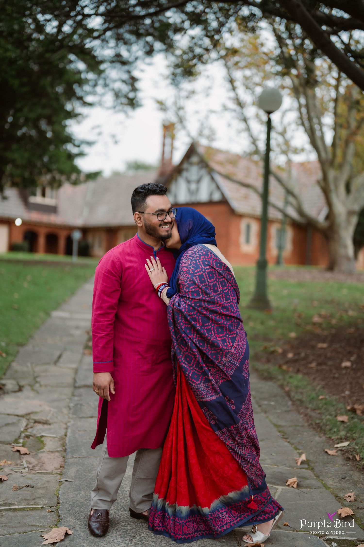 A couple in traditional South Asian attire share an intimate moment on a stone path surrounded by autumn trees.