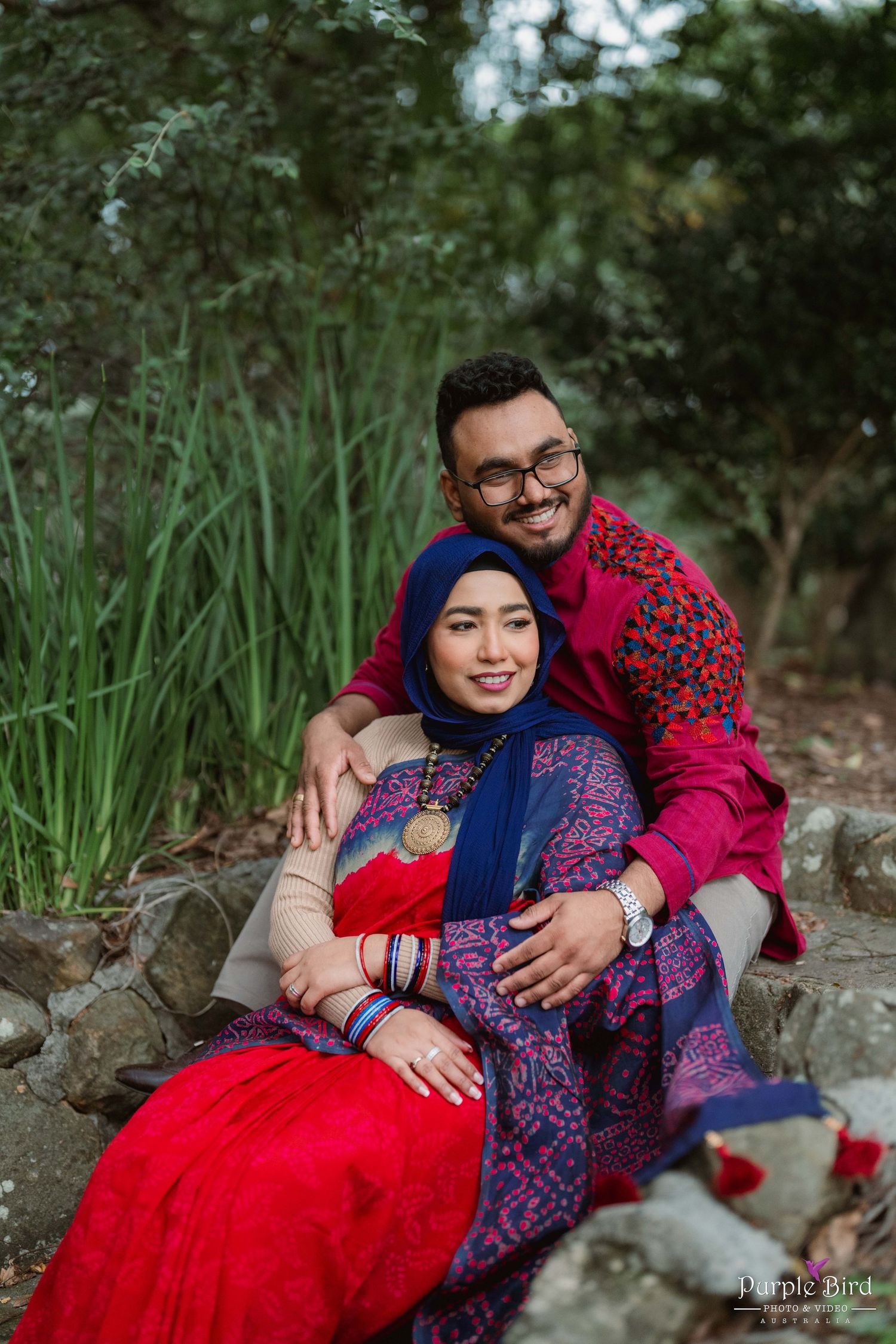 A romantic couple poses together on natural stone seating surrounded by greenery in an outdoor setting.