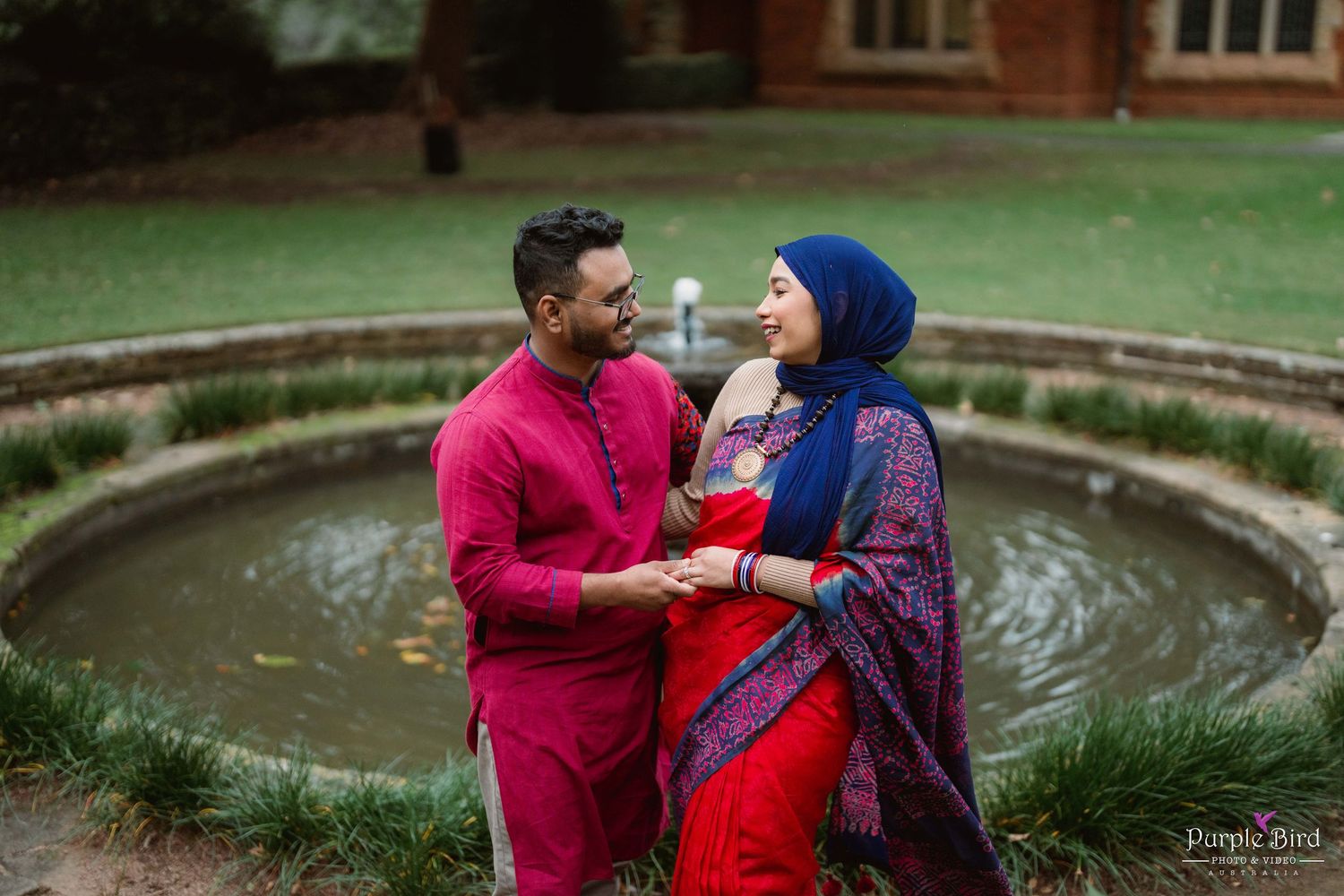 A couple in red and blue outfits share intimate moments in a garden setting during their engagement photoshoot.