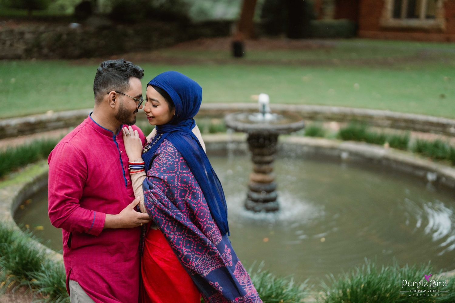 A romantic couple shares an intimate moment in an outdoor garden with a decorative fountain in the background.