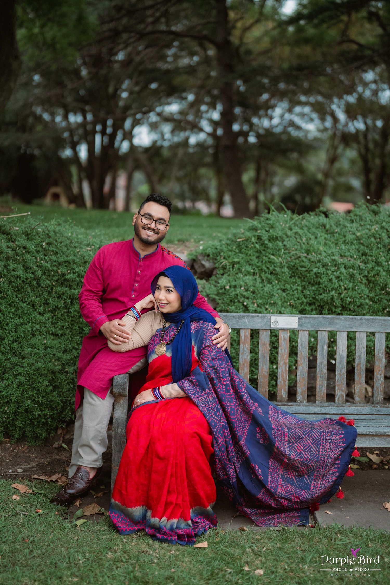 A couple shares an intimate moment relaxing on a park bench surrounded by lush green hedges and trees.