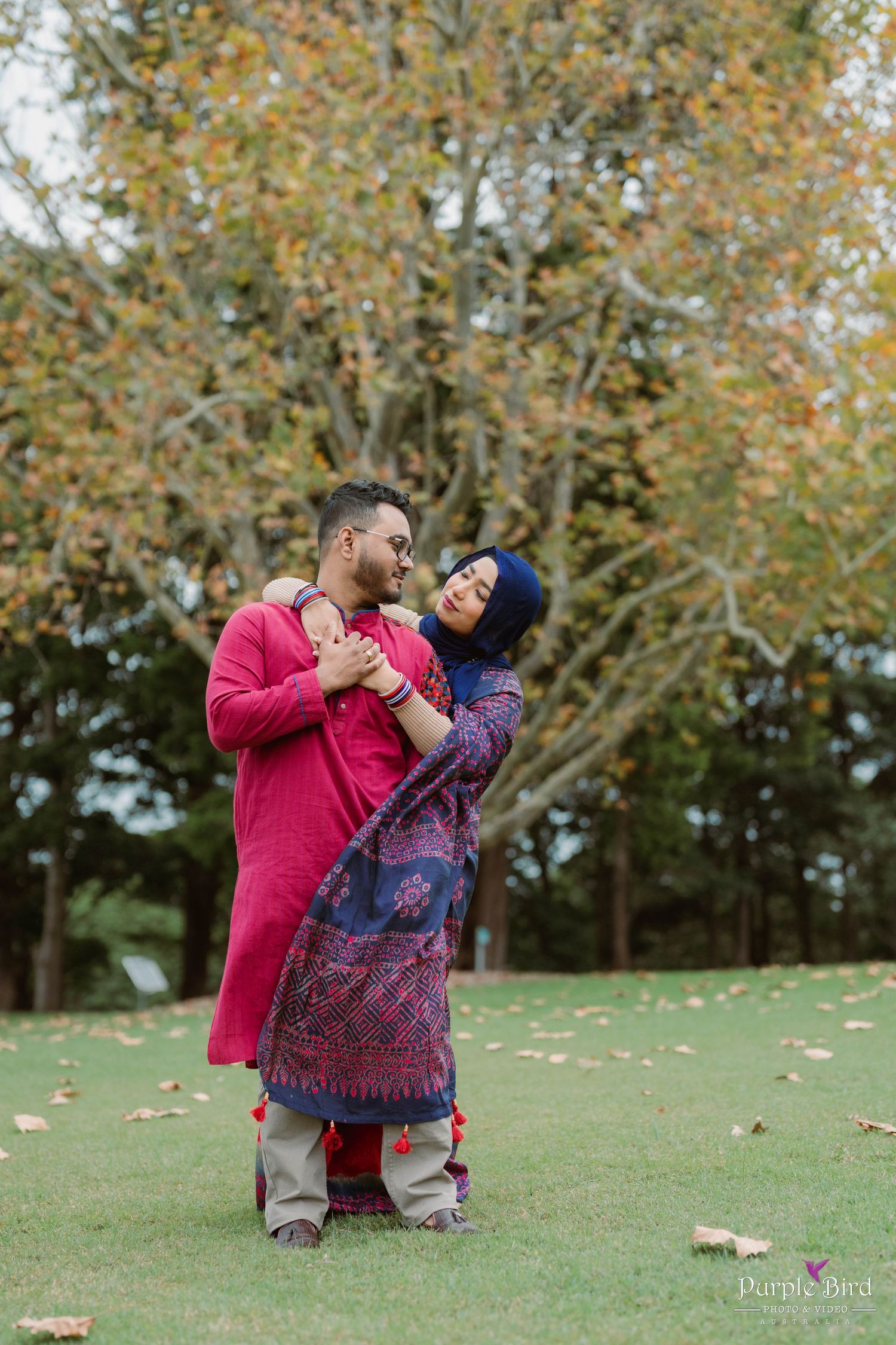 A couple shares a romantic autumn moment together on a grassy hill with fall foliage in the background.
