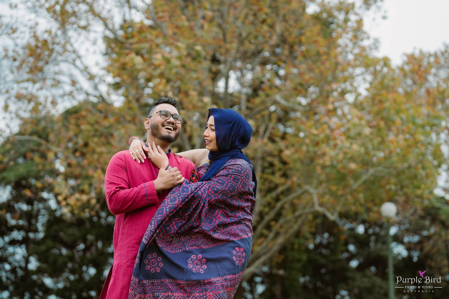 A couple embraces outdoors with autumn trees creating a scenic natural backdrop.