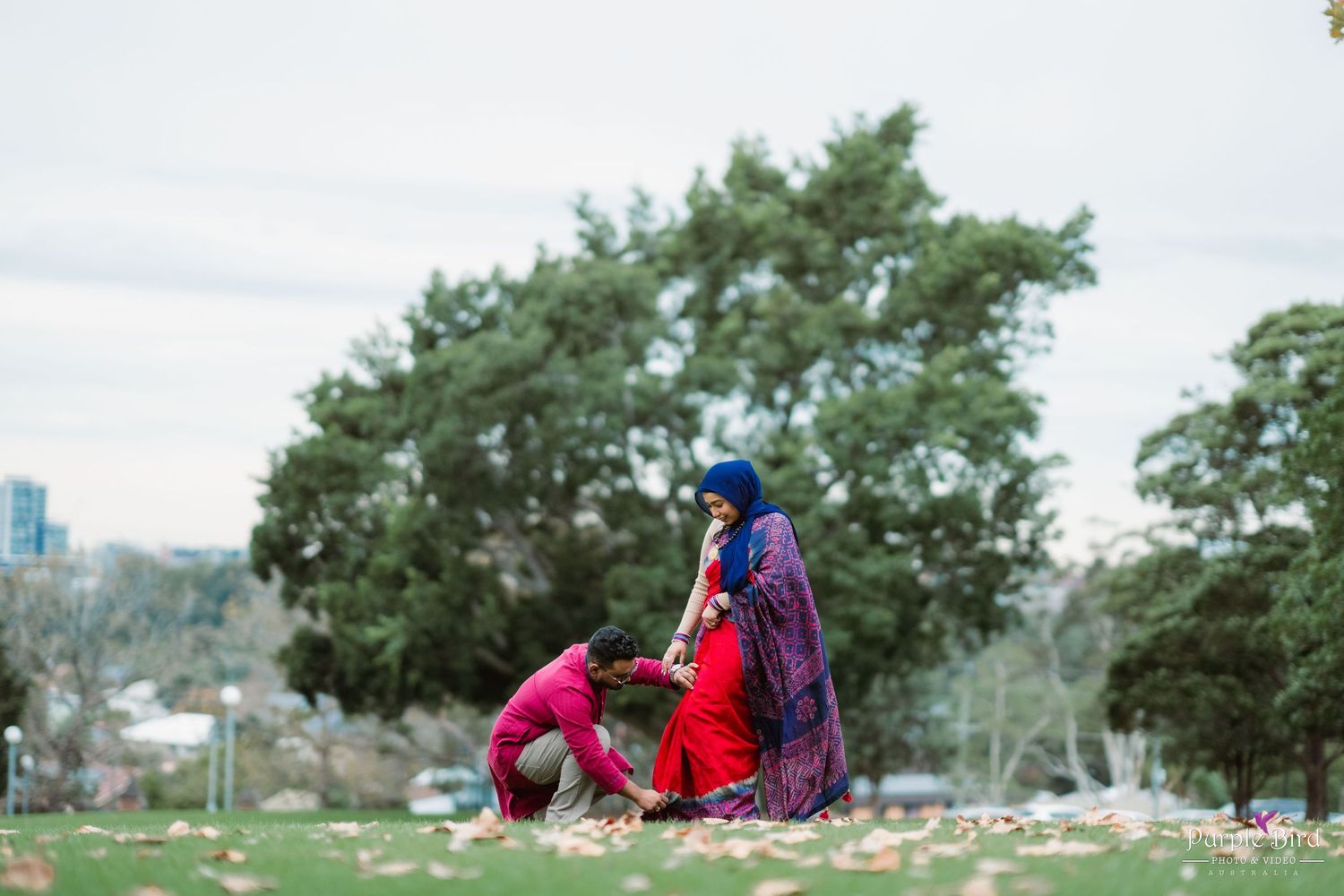 A couple embraces in a green park setting during early spring with trees and grass in the background.