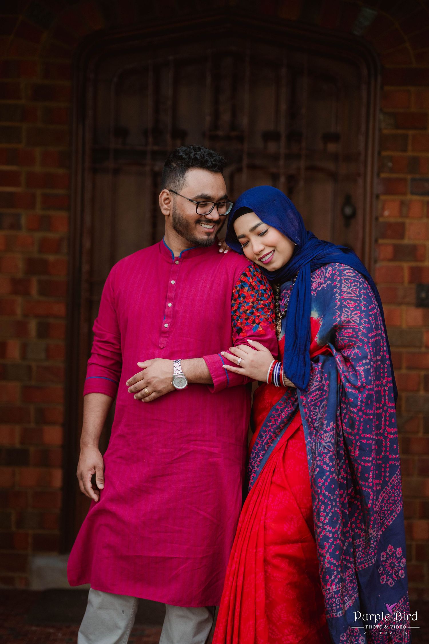 A couple wearing vibrant traditional South Asian attire in bright pink and blue stand affectionately by a brick wall.