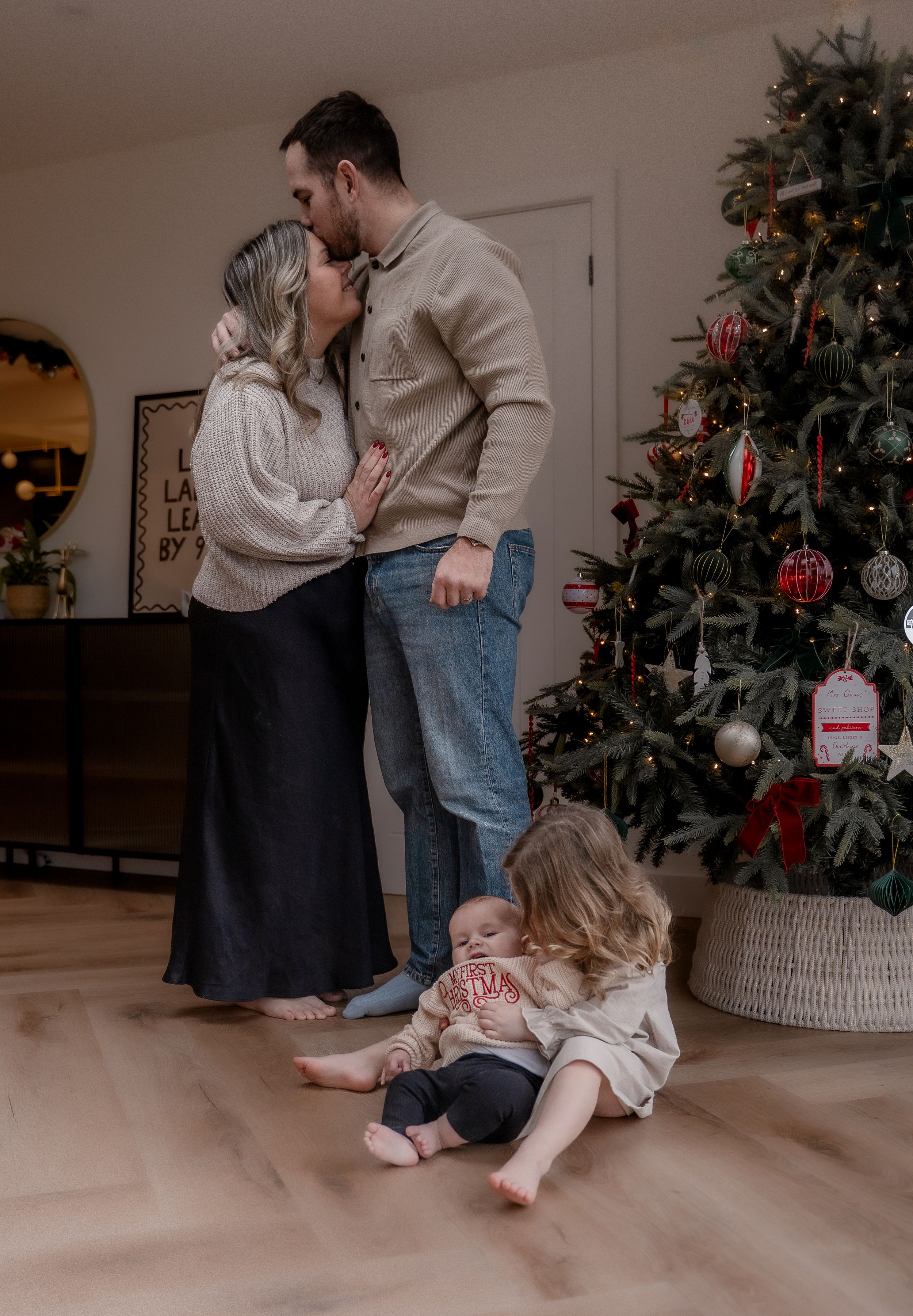 A couple shares an intimate moment by their decorated Christmas tree while their child rests on the floor.