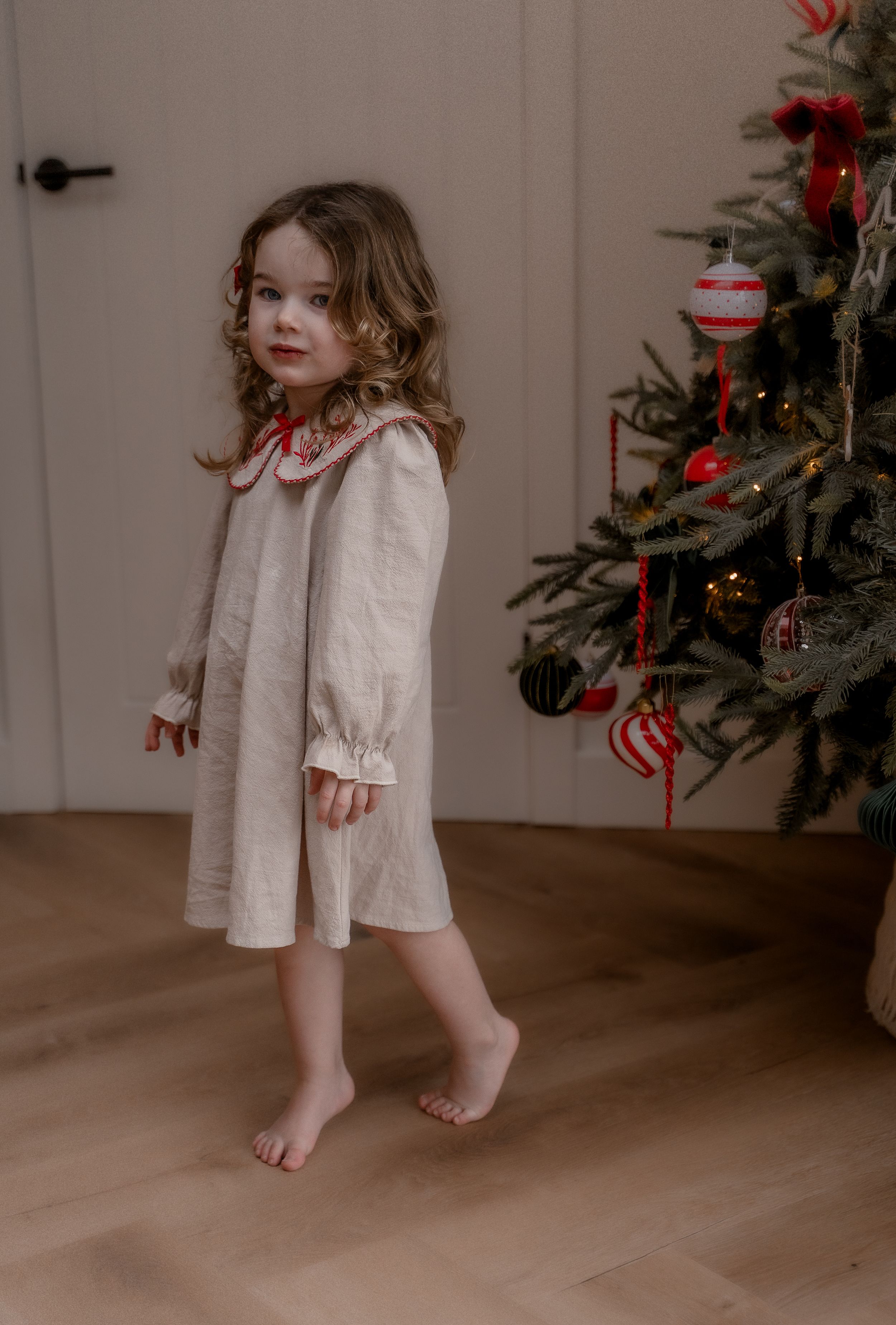 A child in a white dress twirls and dances near a decorated Christmas tree in a sequence of candid holiday photos.