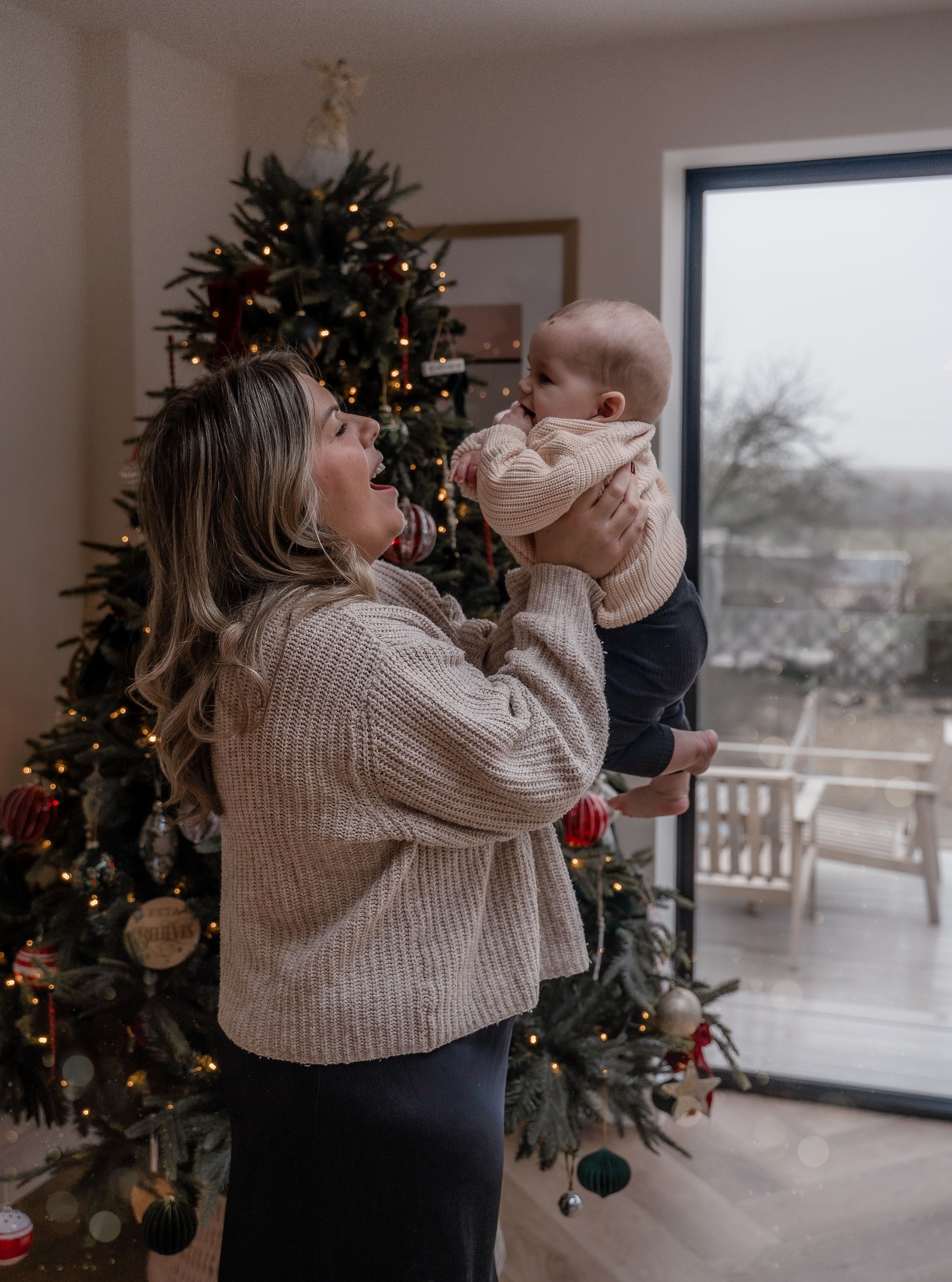 A cozy holiday scene with a decorated Christmas tree, a beige sweater, and a baby being lifted up in a modern living room.