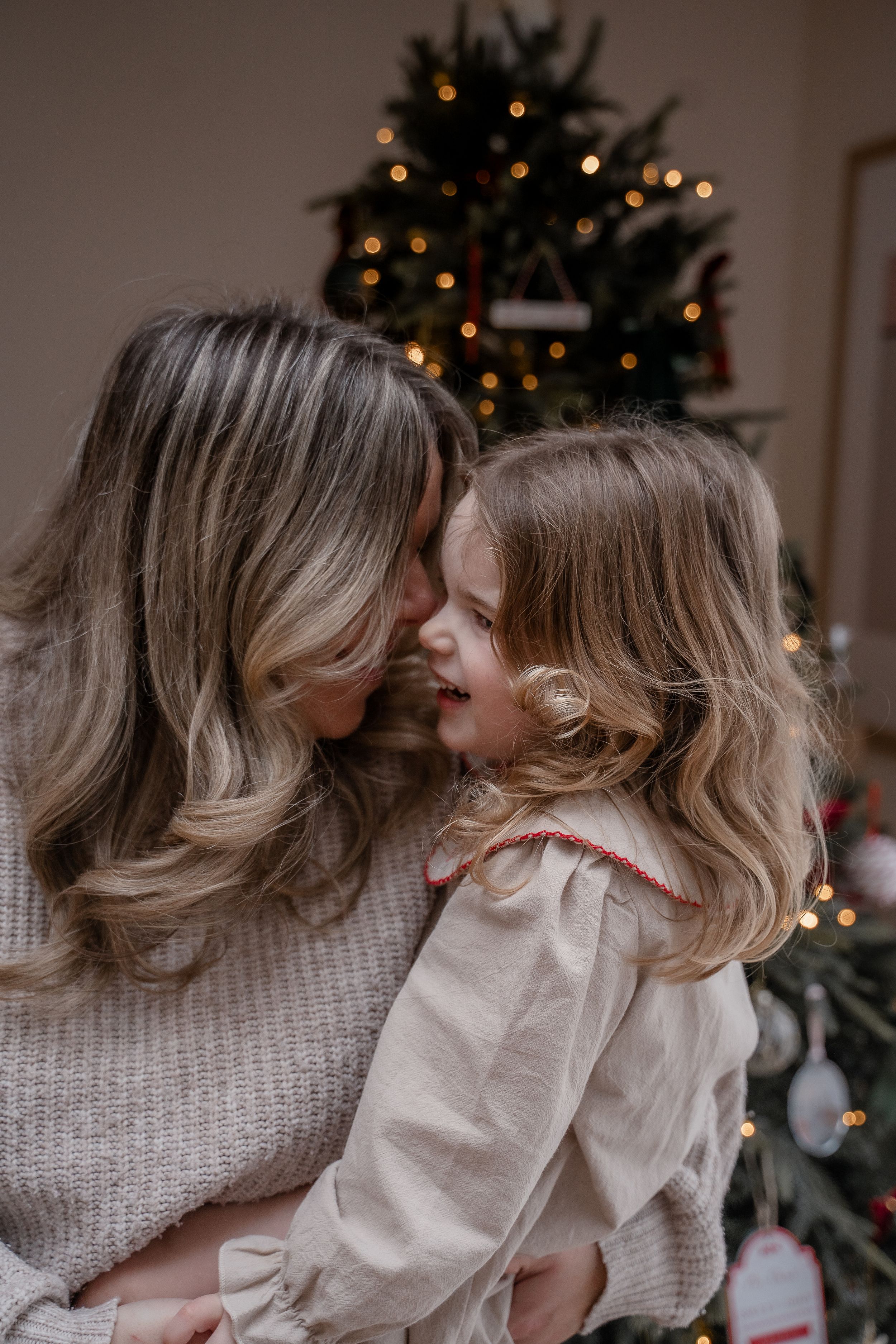 Mother and daughter share tender moments in front of a Christmas tree with twinkling lights in the background.