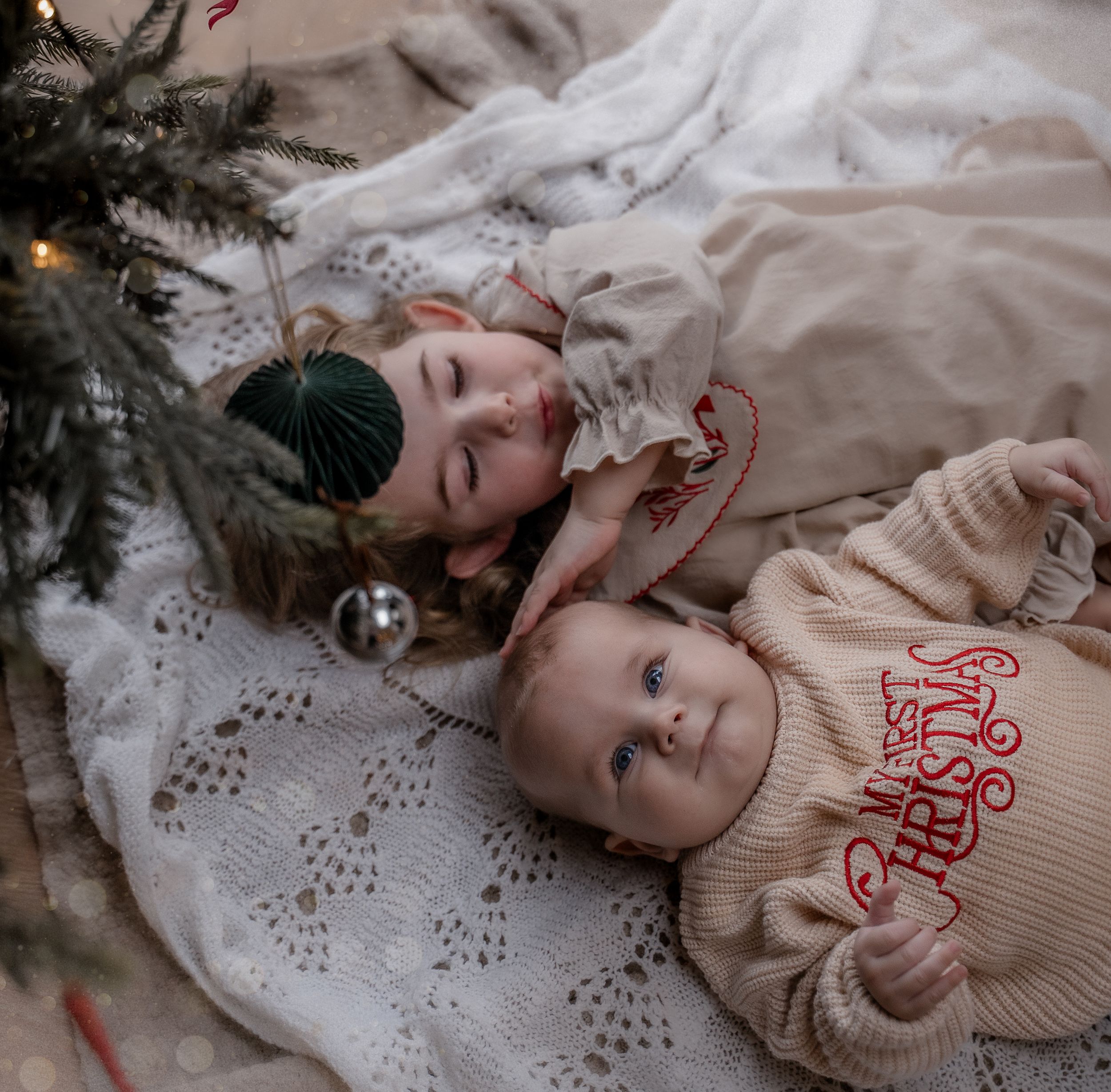 Two children lying on a cozy white blanket next to a Christmas tree, wearing festive winter sweaters.
