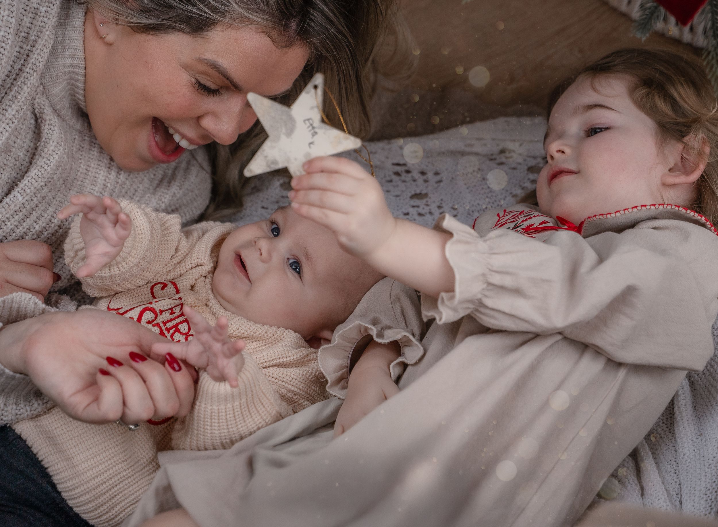 a mum lying down under a christas tree with her daughter and baby son smiling
