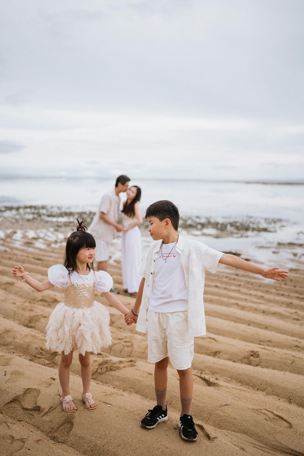 A family dressed in white outfits poses together on a sandy beach during an overcast day.