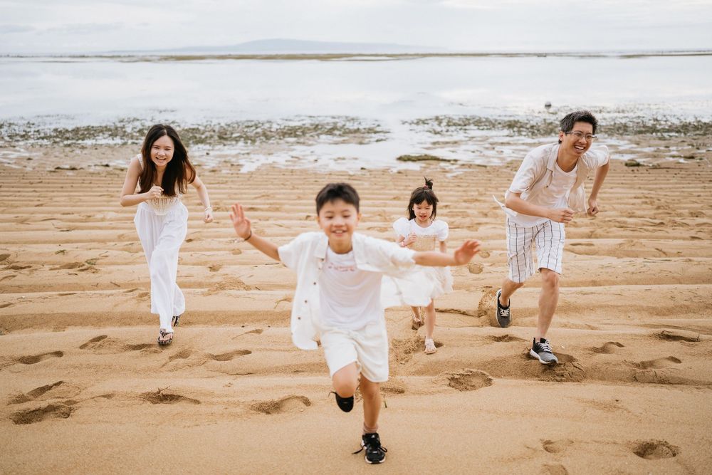 A family in white clothing runs playfully across a beach with the ocean in the background.