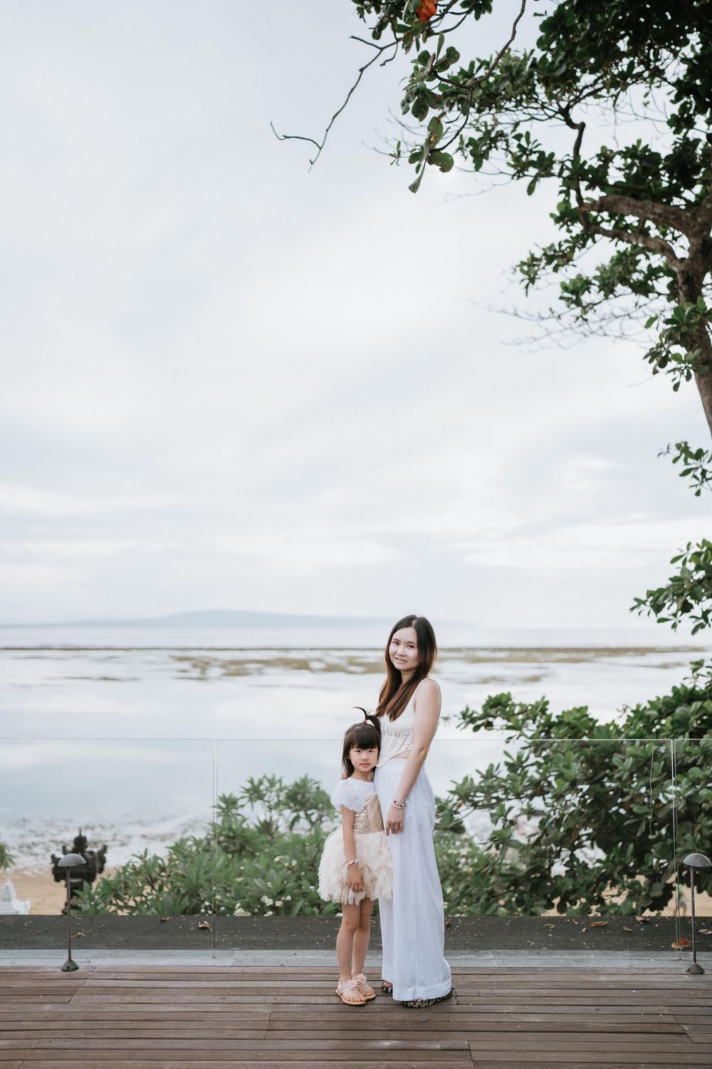Two people in white outfits stand on a wooden deck overlooking a scenic coastal view.