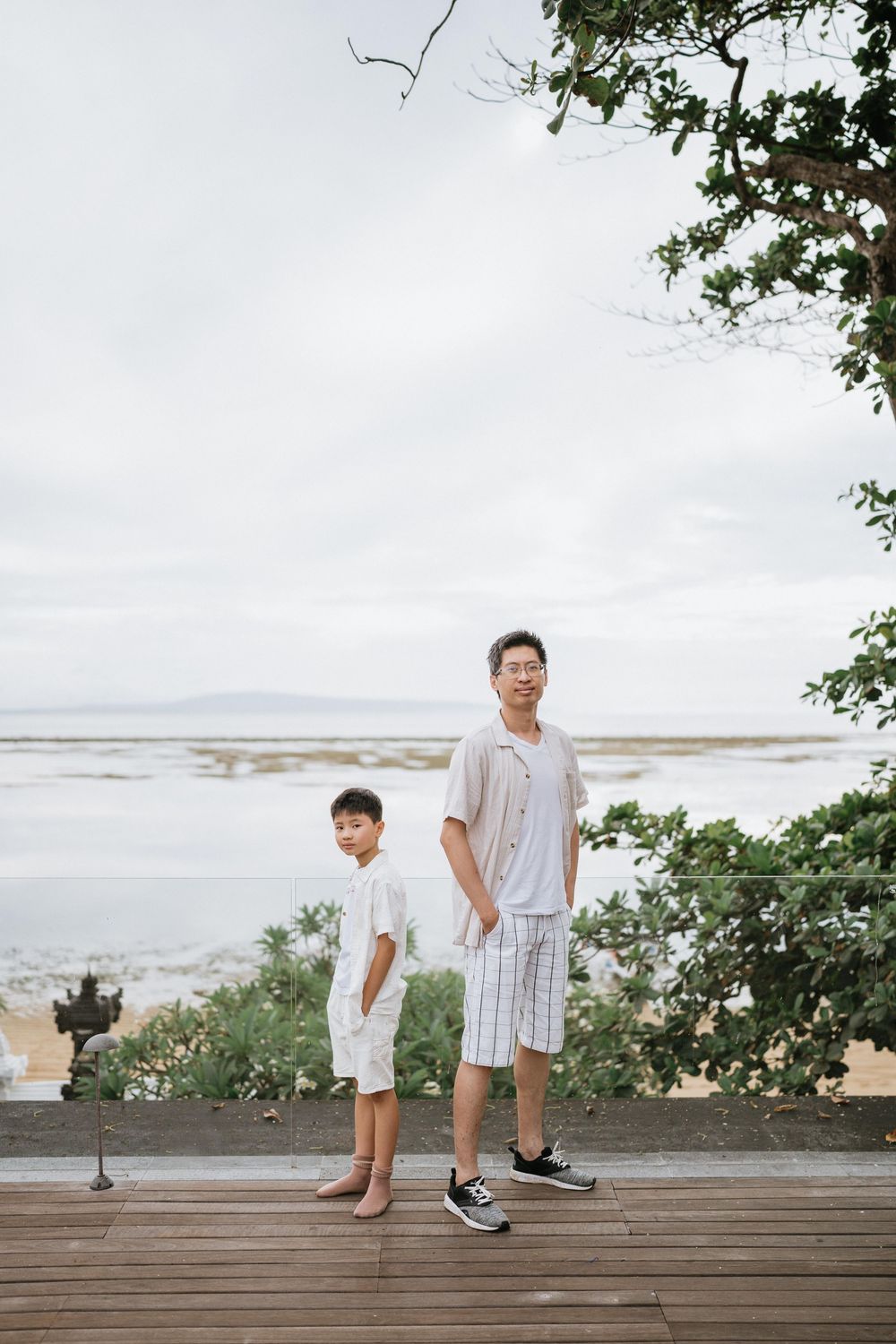Two people in light colored clothing pose on a wooden deck with an ocean view.