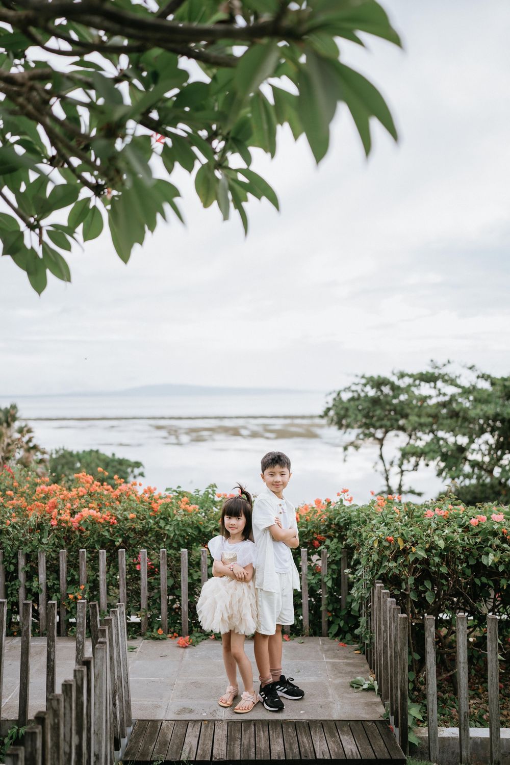 Two children in white outfits stand on a wooden walkway surrounded by orange flowers and coastal views.