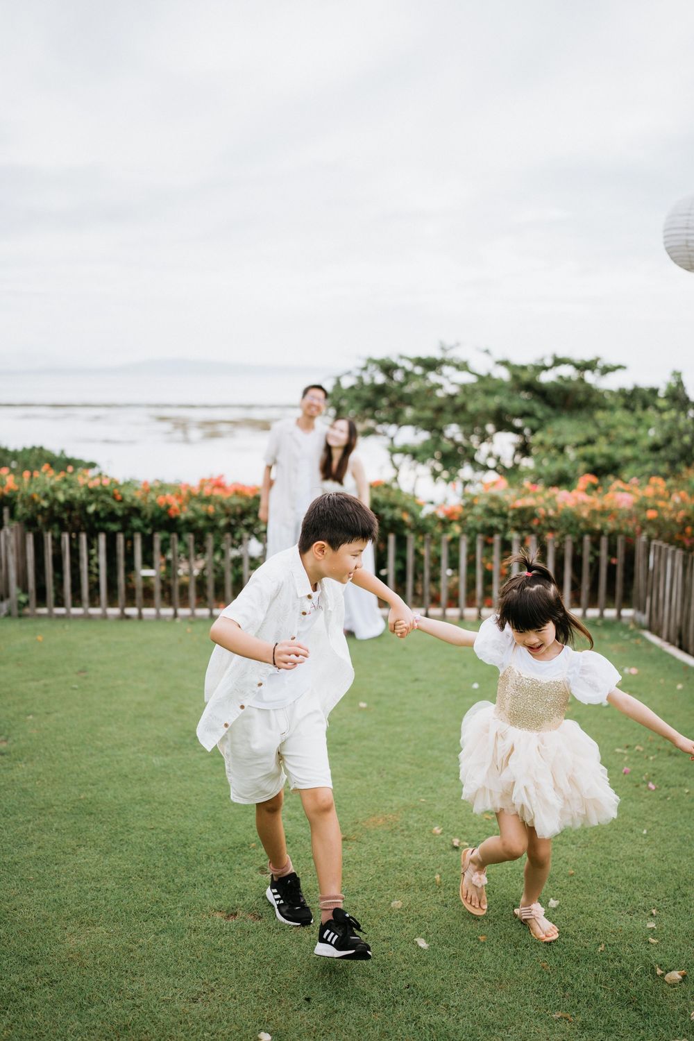 Children in white outfits play on a grassy area with orange flowers and ocean views in the background.