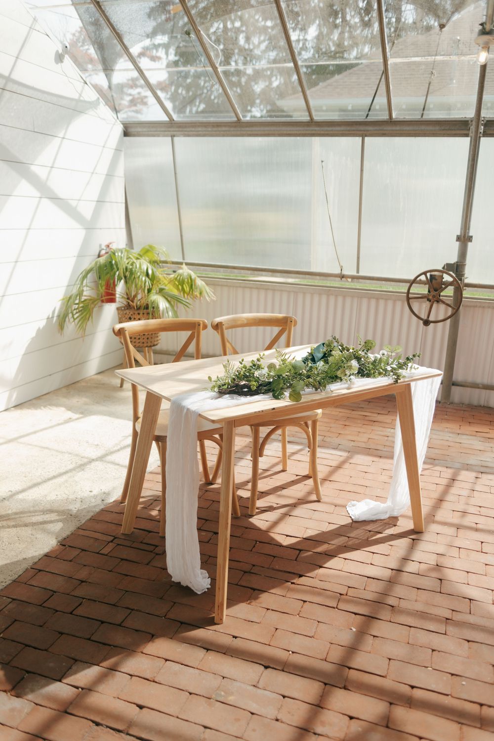 Rustic wooden table decorated with greenery and cloth runner in a bright sunlit greenhouse interior.