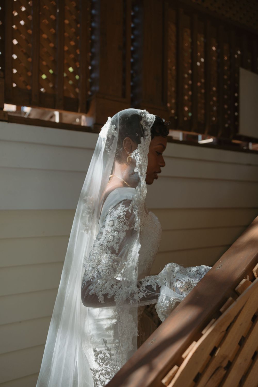 A bride in a lace wedding dress and long veil stands next to wooden chairs during a photoshoot.