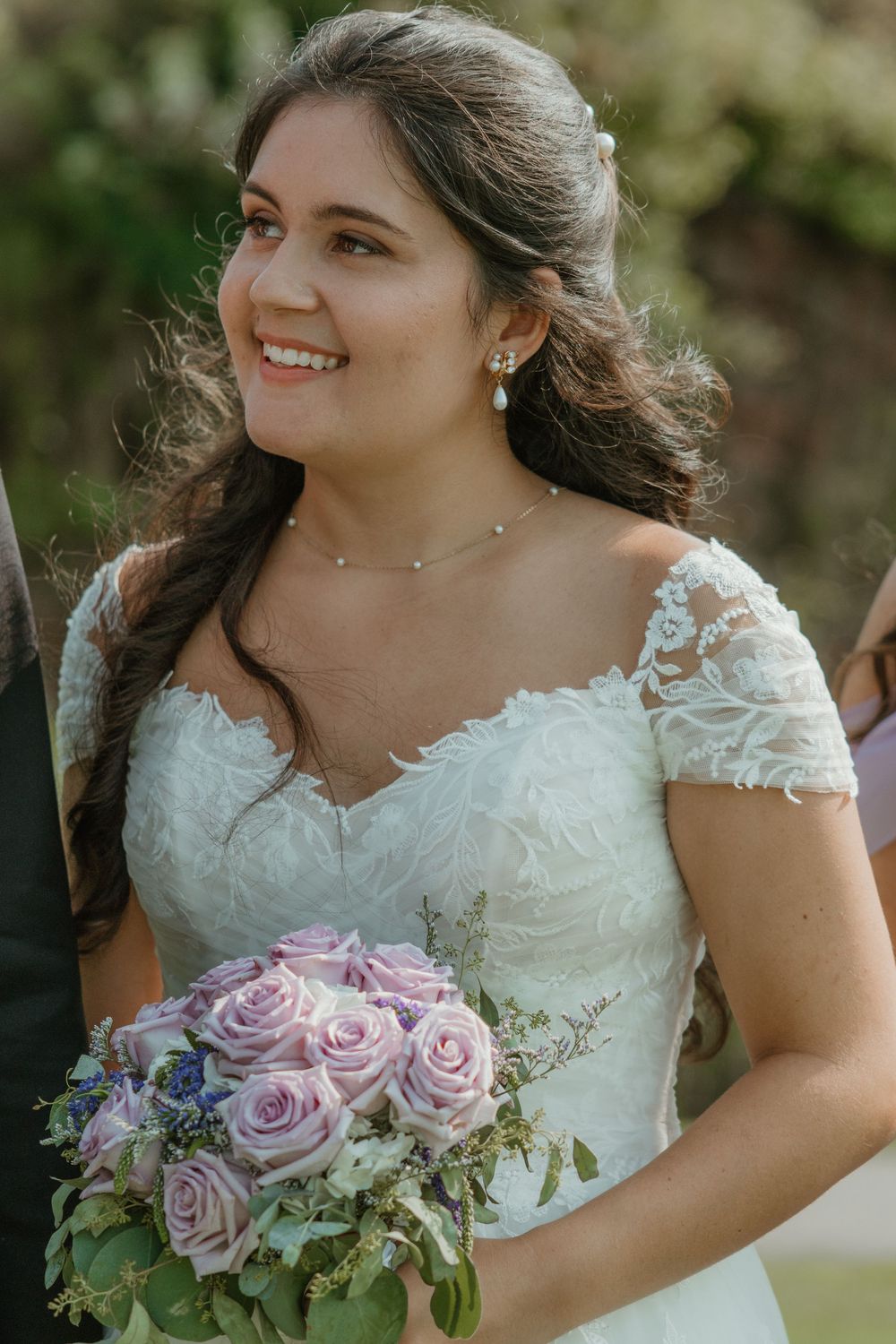 Bride in lace dress holds a beautiful bouquet of lavender roses and greenery.