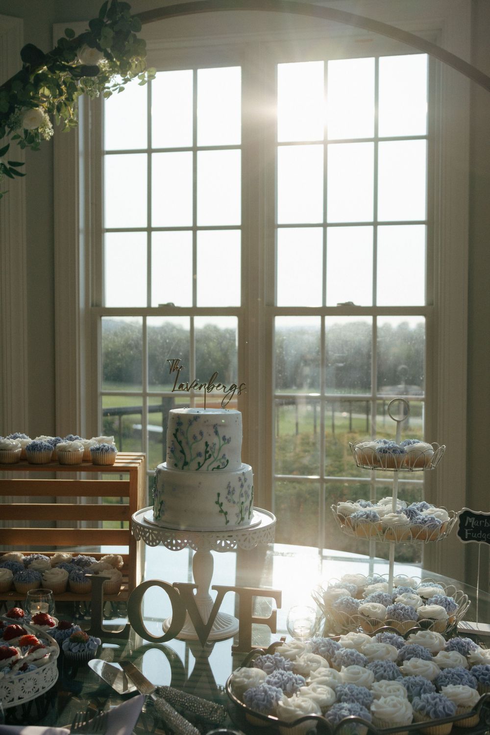 Wedding dessert table layout with cupcakes and cake stand displayed in front of bright sunlit windows overlooking scenic views.